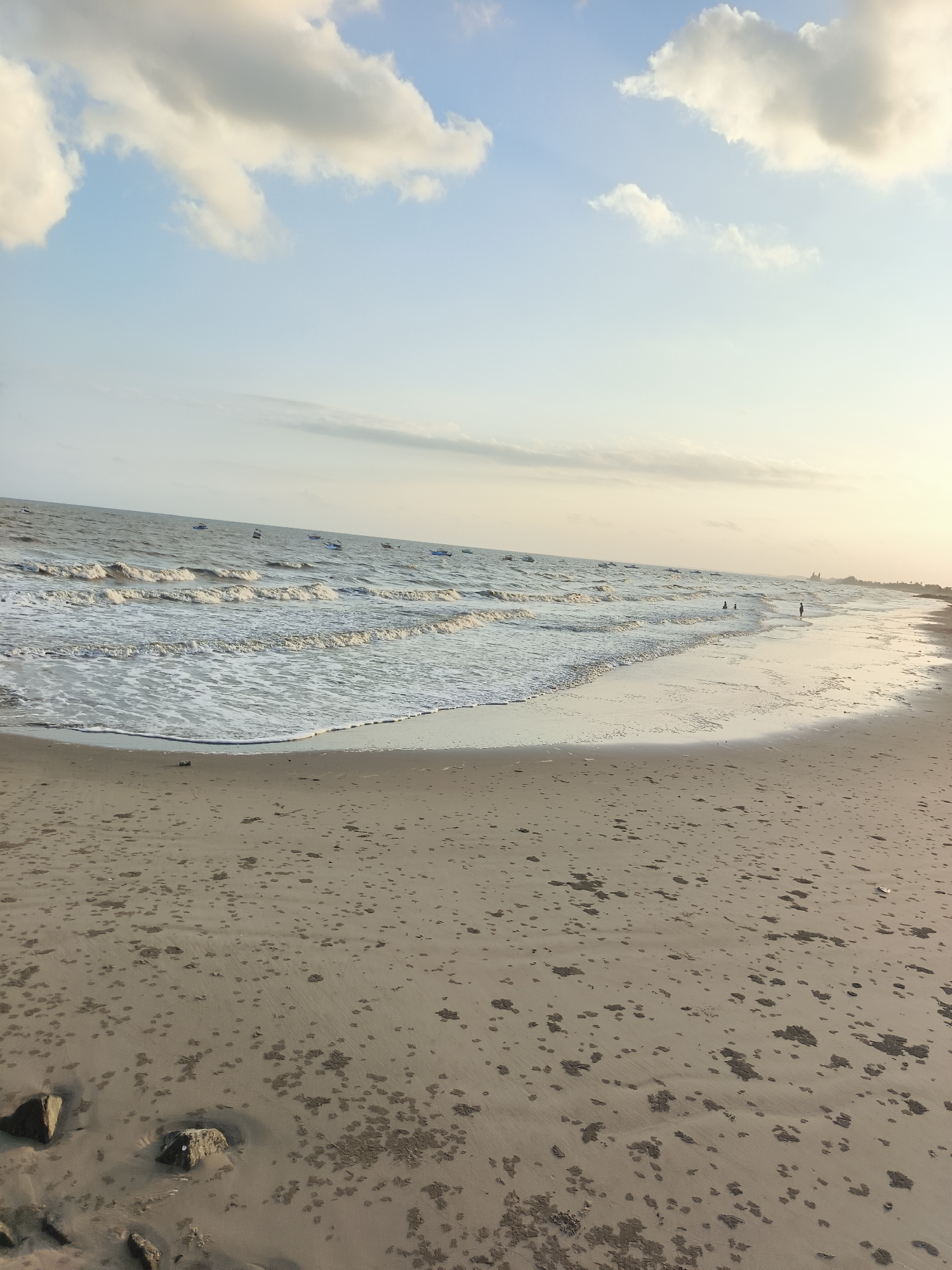 The image depicts a tranquil beach scene at Kupang Berkah Jaya, Indonesia, during what appears to be late afternoon or early evening. The sky is a gradient of soft blue and pale orange, with scattered white and yellowish clouds. The ocean stretches across the frame, with gentle waves rolling towards the shore, creating white foam. Several small boats are visible scattered across the water. On the beach, a few individuals are present: two people are partially submerged in the water, and one person stands on the wet sand near the shoreline. The beach itself is composed of light brown sand, marked with small dark patches that could be seaweed, debris, or tide marks. A few rocks are visible in the foreground. The overall impression is one of peacefulness and natural beauty.