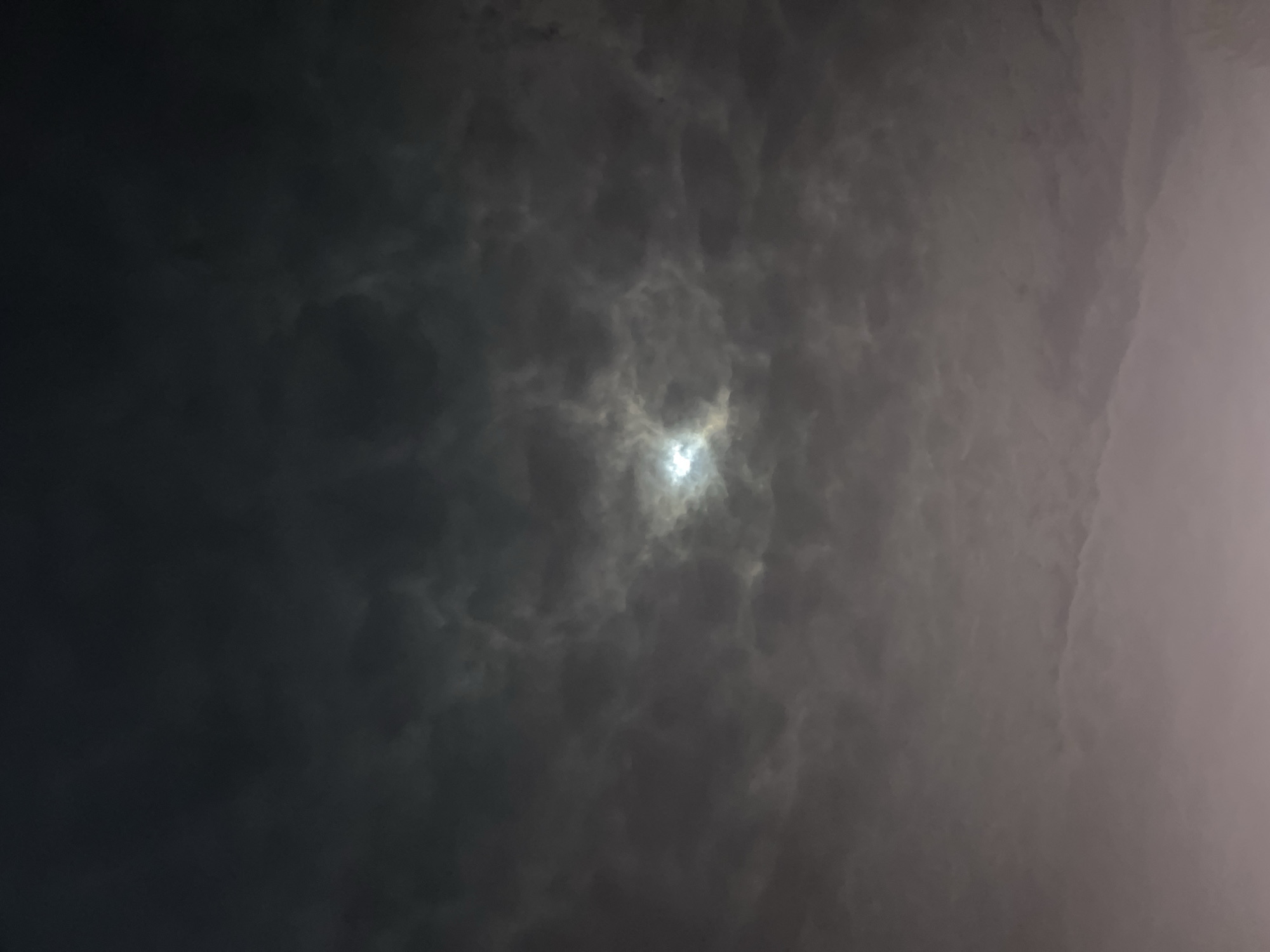 The image presents a heavily overcast sky, filled with dense, dark clouds. A prominent, diffused bright light source is visible through a break or thinner section of the clouds, positioned slightly above the center of the frame. This light illuminates the surrounding cloud formations, creating a radiant glow that contrasts with the deeper grey and black tones of the denser cloud masses, especially towards the left and upper left. The clouds exhibit significant texture and depth.
The overall low light levels suggest a nighttime scene with the moon behind the clouds, or a heavily overcast day at dawn or dusk where the sun is almost completely obscured. There are no visible people, objects, or specific activities taking place, as the image focuses solely on the sky. The weather is clearly cloudy. No discernible location cues for Kotel, Bulgaria, are present within the visual content of the image itself, and there is no visible text.