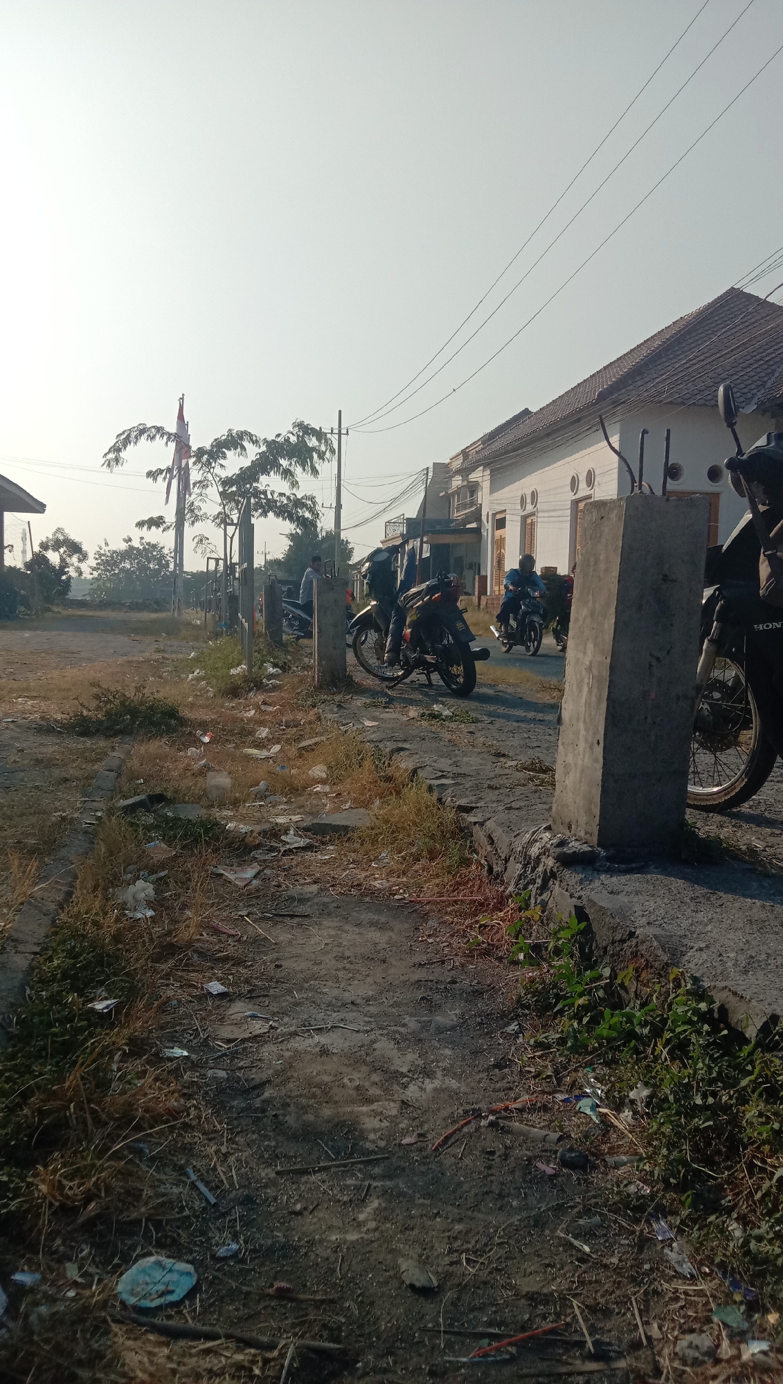 The image depicts a street scene in Curah Dukuh, Indonesia. In the foreground, a rough, unpaved path is visible, littered with dry grass and various pieces of trash, including plastic wrappers and debris. To the right, a concrete structure, possibly a barrier or foundation, stands prominently, with a motorcycle partially visible behind it. The path leads towards a paved road where several motorcycles are parked or in motion. In the background, a white building with a tiled roof lines the street. A flagpole with the Indonesian flag can be seen to the left. Several individuals are present, some standing near motorcycles and others riding them. The sky is clear and bright, suggesting daytime.