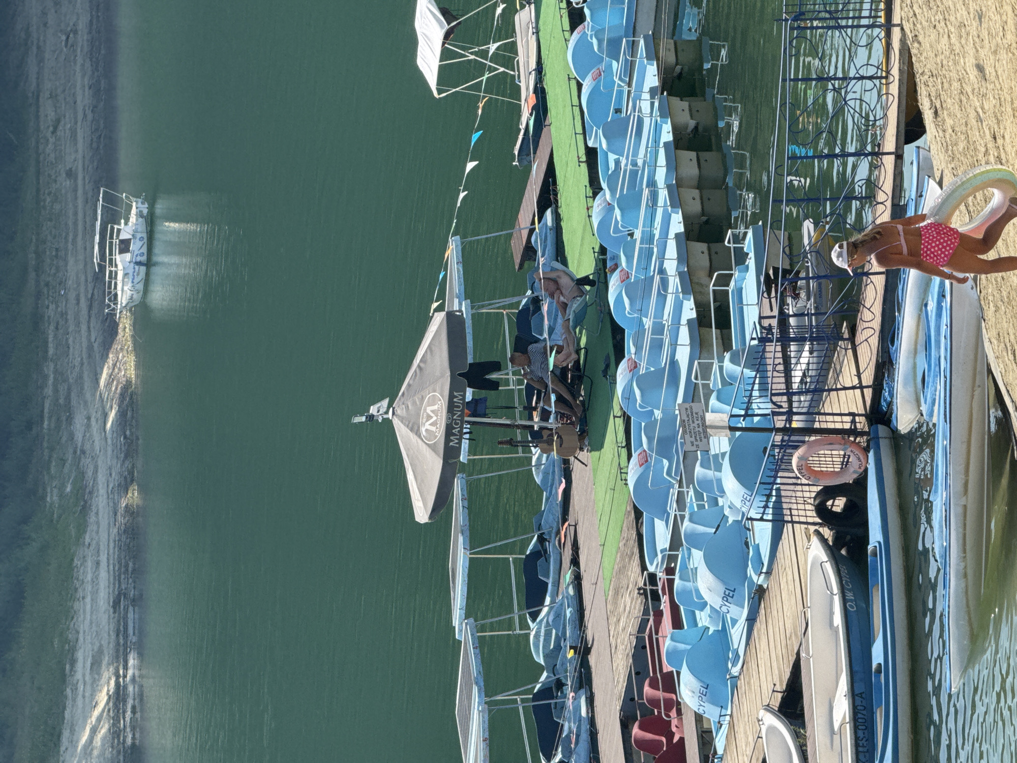 The image depicts a sunny day at a water recreation area in Polańczyk, Poland, likely on the Solina Lake. In the foreground, a young girl in a pink polka-dot bikini walks along a sandy shore, carrying a colorful inflatable ring. Several boats are docked, including a blue and white motorboat with a registration plate visible. A wooden pier extends into the water, lined with numerous blue pedal boats, some with canopies. On the pier, under a large umbrella bearing the "Magnum" logo, two men are relaxing. One man is lying on a lounge chair, while the other is seated. Behind them, a guitar rests against a support. Further out on the water, a small white motorboat is anchored near the shore, which is covered in light-colored sand and sparse vegetation. The water is a deep green, reflecting the clear sky and surrounding landscape.