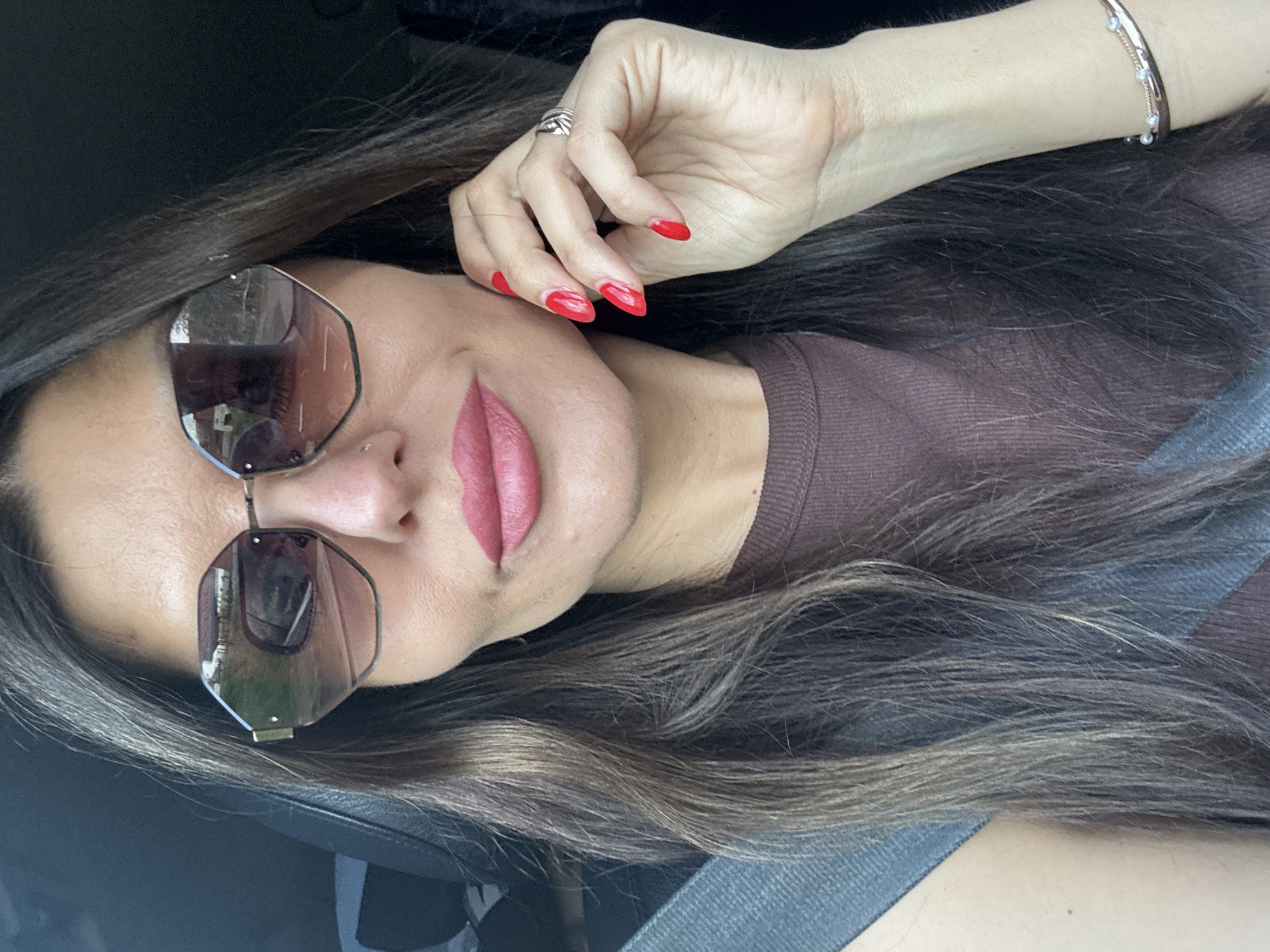 In this close-up, forward-facing selfie taken in the city of Chaikovsky, Russia, a woman is seen wearing large, hexagonal sunglasses with a gradient lens, reflecting a street scene with buildings and trees. She has dark, long hair, a nose piercing, and her lips are painted with a matte pink lipstick. Her right hand, adorned with bright red nail polish and a silver ring, is raised to her cheek. She is wearing a brown ribbed top and a seatbelt is visible across her chest, indicating she is likely in a car. A thin bracelet with small pearls is visible on her right wrist. The background is dark and out of focus, suggesting the interior of a vehicle.