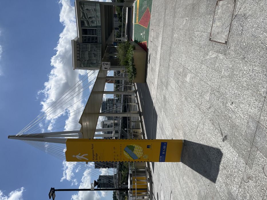 The image captures a vibrant, sunny day in a modern urban pedestrian area of Brisbane City, Australia. The central feature is the sleek, white cable-stayed Kangaroo Point Bridge, which dominates the left side of the frame with its elegant pylon and numerous tensioned cables stretching into a bright blue sky dotted with fluffy white cumulus clouds.
In the foreground, a prominent yellow information sign stands vertically on the paved walkway. The sign clearly displays the text "Kangaroo Point Bridge" along with a pedestrian symbol and a map of the surrounding area, indicating the bridge spans a body of water, likely the Brisbane River. The ground is paved with light grey, textured rectangular tiles, reflecting the strong sunlight. Shadows cast by the sign and nearby structures suggest it is mid-morning or early afternoon.
Modern multi-story buildings with reflective glass facades are visible in the background, appearing both behind and beneath the bridge's structure. A small planter box with green foliage adds a touch of nature near one of these buildings. A white sign with symbols for both pedestrians and cyclists is visible further along the path, indicating shared usage. No people are visible, giving the scene a serene, open feel, but the infrastructure suggests it is a popular route for walking and cycling. The overall impression is one of a clean, well-maintained, and visually striking urban landscape.