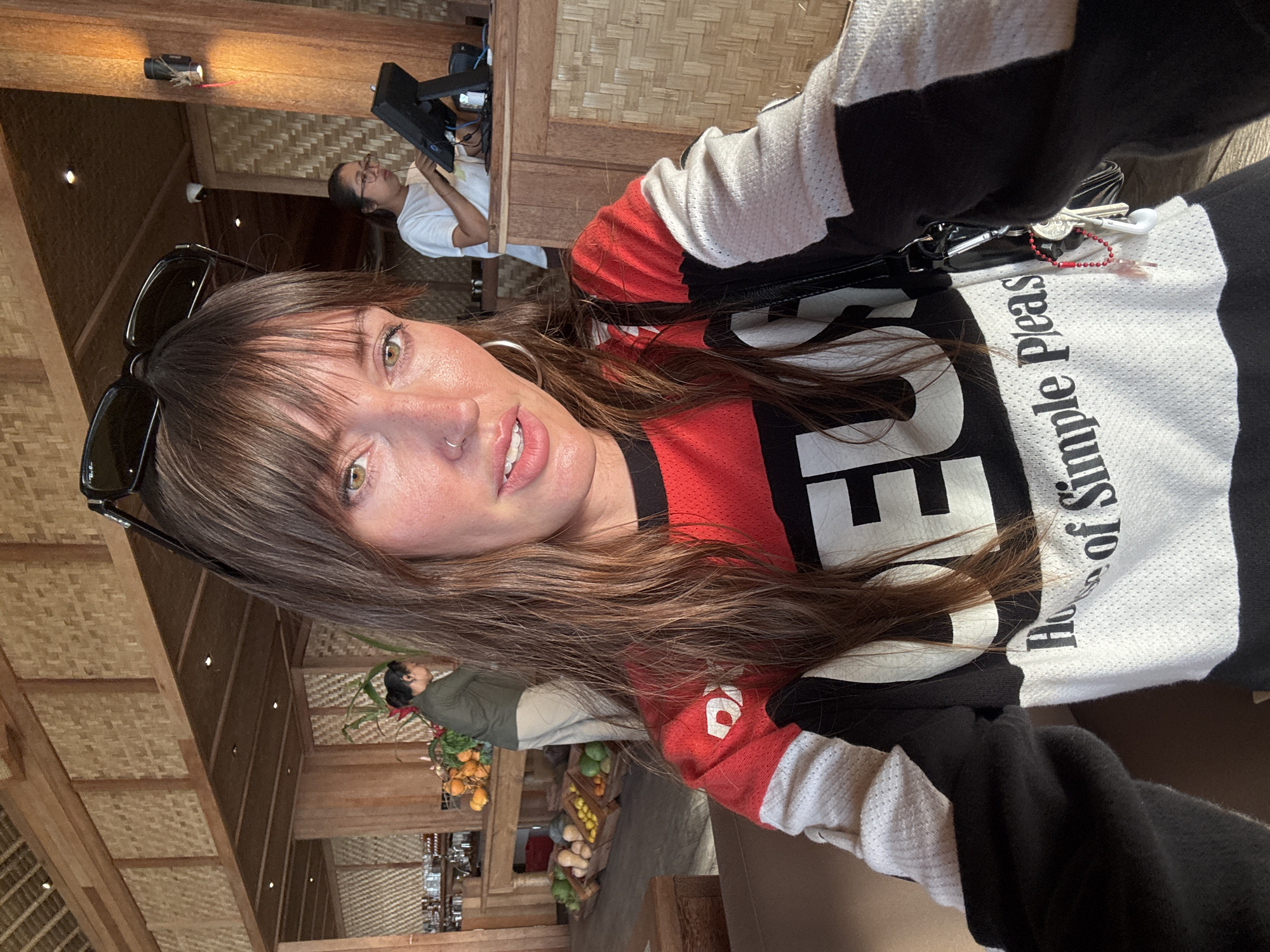 A medium shot shows a fair-skinned woman with long, brown, wavy hair, bangs, and a nose ring posing for a selfie. She is wearing a red, white, and black long-sleeved shirt with the word "DEUS" prominently displayed across the chest in large white letters. Her left hand is visible, holding a black purse that is adorned with keys and a red beaded lanyard. Sunglasses are perched on top of her head. Her makeup is visible, including eyeliner and lip gloss.
In the background, the interior of a restaurant is depicted. The architecture features bamboo walls and wooden accents. To the left, a display of fruit, including oranges and green gourds, is arranged on a wooden table. A person wearing a green top and beige pants is standing behind the table, facing away from the camera. Further back, another person, wearing glasses and a white t-shirt, is visible at a counter, interacting with a POS system. The environment suggests a casual dining setting, possibly a resort or cafe, located in Kuta Selatan, Indonesia, given the regional architectural style and commonality of such establishments. The lighting is bright and natural, indicating daytime.