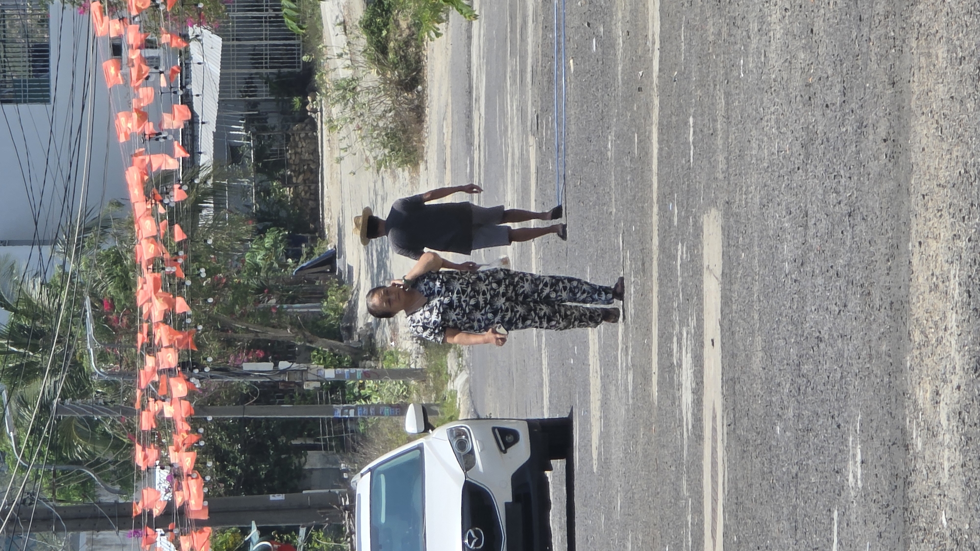 The image shows a street scene in Bắc Nha Trang, Vietnam. Two individuals are walking on a paved road with white lane markings. One person, wearing a dark gray shirt and shorts, has a straw hat. The other person wears a patterned, dark-colored, short-sleeved pajama set. A white Mazda SUV is parked on the left side of the road, facing the viewer. Overhead, a string of small orange flags with a central symbol (possibly communist symbols) spans the road, connected to buildings and poles. Vegetation and buildings are visible on the sides of the road. The sky is not visible.