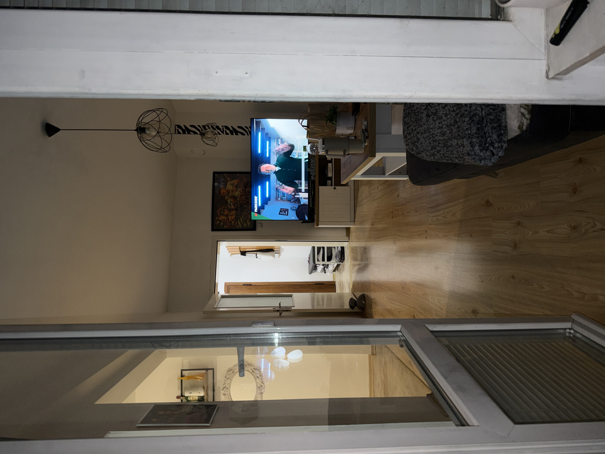 This image captures an indoor residential scene, likely an apartment or house, viewed from a threshold looking down a hallway into a living area.
In the immediate foreground on the left, a white PVC-framed glass door or window is ajar. Its glass pane reflects elements from an adjacent room, including a framed picture, a wall-mounted shelf holding two bottles and a yellow object, and an ornate white oval mirror. The lower section of this door/window features horizontal internal blinds.
The central view extends down a hallway with light-colored wooden laminate flooring. Midway down the hallway on the right, a small grey stuffed animal sits on the floor. The hallway culminates in an open doorway. Through this doorway, a dark wooden coat rack with a white bag and a dark garment hanging on it is visible against the far wall. Below the coat rack, a grey metal shoe rack displays multiple pairs of mostly white sneakers. A dark wooden door frame outlines the entry to this further room.
To the right of the hallway, a living room is visible. A large flat-screen television is centered on a light-colored wooden and white media console. The TV displays a man with dark hair, wearing a dark green shirt, gesturing with both hands. A "PARKSIDE" logo is visible in the upper left corner of the screen. Above the television, a dark-framed picture or artwork depicting multiple figures hangs on the wall. To the right of the TV, a wall section is adorned with a black and white zebra-striped pattern. A modern pendant light with a black wire framework hangs from the white ceiling in this area.
In front of the television, a light-colored wooden coffee table with white legs holds several items: a large grey insulated mug, a clear plastic water bottle, a small white potted plant, and a small dark object (possibly a lighter or electronic device). Partially visible behind the coffee table is a dark sofa covered with a grey, textured blanket. On the far right, a white window frame with blinds is partially visible. The scene is illuminated by interior artificial lighting, creating soft shadows across the wooden floor.