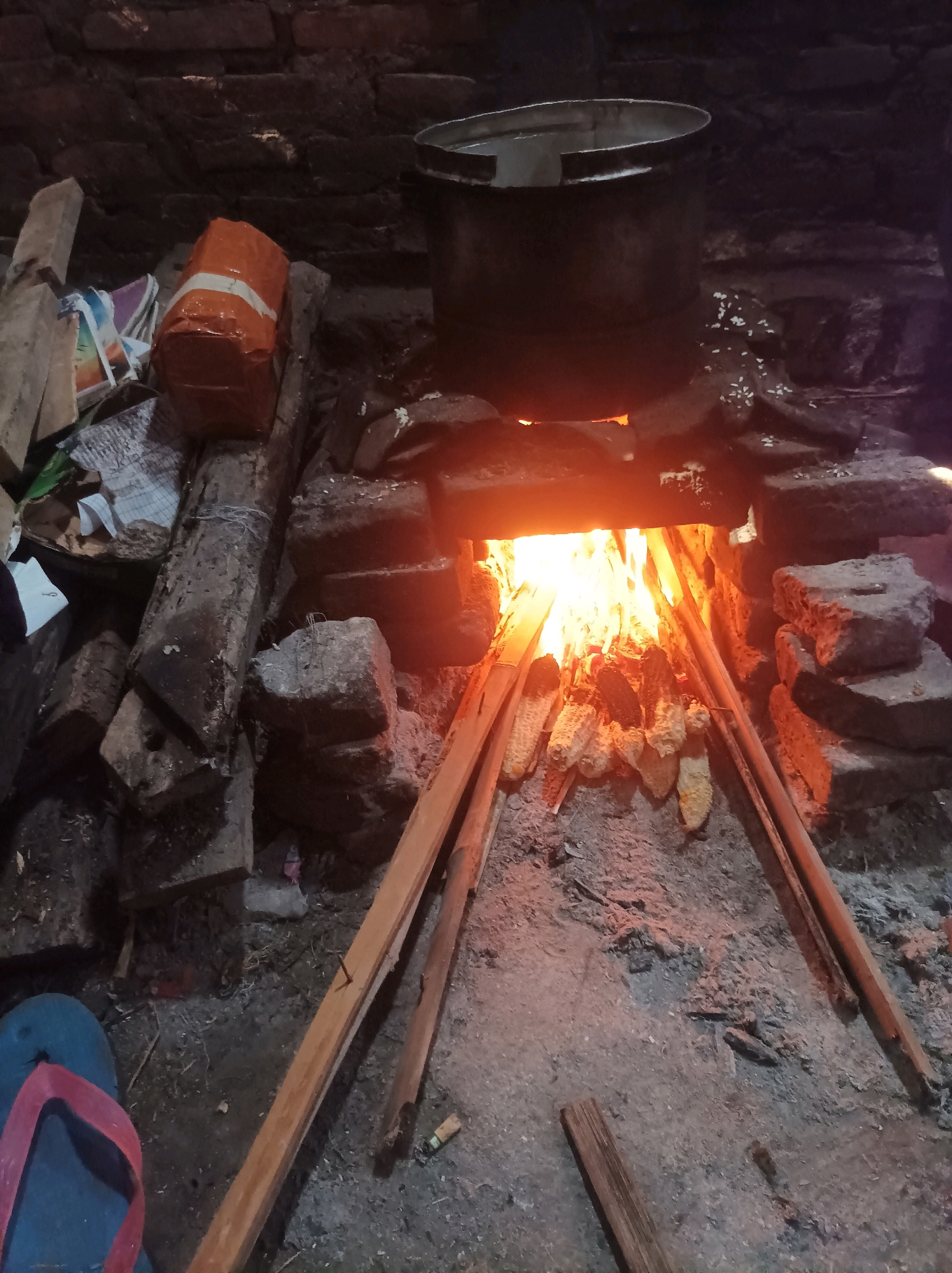 The image depicts a rustic outdoor cooking setup, likely in Canga'an, Indonesia. A fire burns brightly in a makeshift brick oven, illuminating several ears of corn being roasted over the flames. Wooden sticks prop up the corn, and long wooden planks are arranged in the foreground. To the left, a bundle wrapped in orange paper and white tape rests on some logs and what appear to be newspapers. A metal pot sits atop the oven, suggesting it is also being used for cooking. The ground is covered in dirt and ash. In the lower left corner, a blue and red flip-flop is visible. The overall environment suggests a simple, traditional method of food preparation.