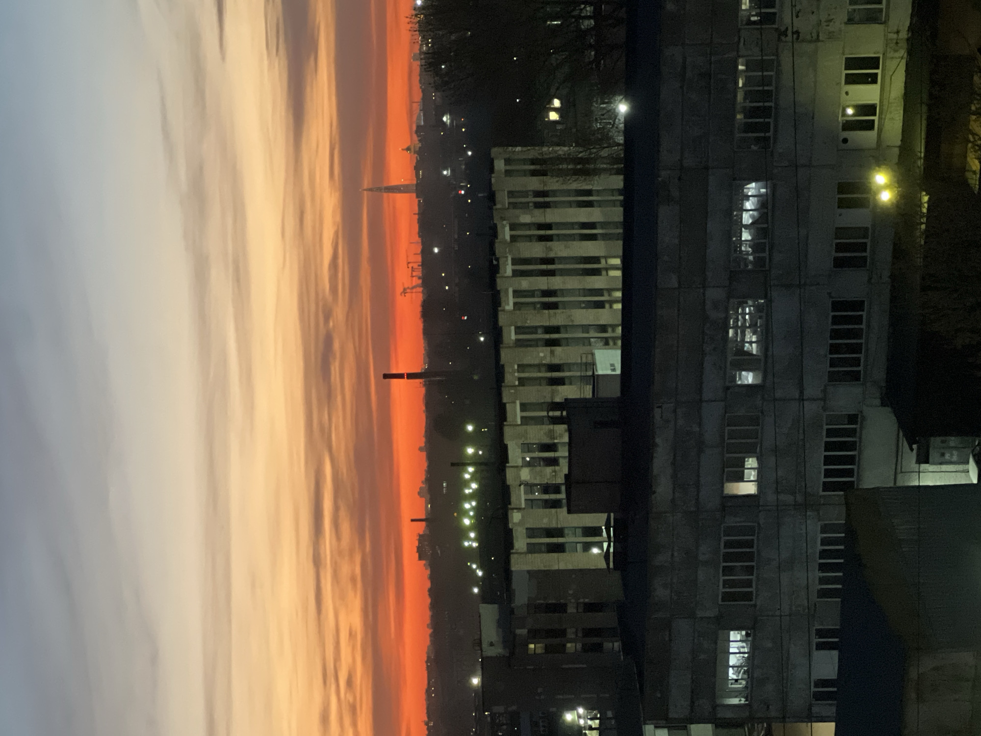 An urban landscape is captured during either sunrise or sunset, viewed from an elevated perspective. The sky exhibits a striking gradient of colors, transitioning from deep red and vivid orange at the horizon to lighter orange, yellow, and finally light grey and blue towards the top. Streaky clouds are present across the colorful expanse.
Along the horizon, a distant city skyline is silhouetted, featuring several chimneys or smokestacks and numerous scattered artificial lights from buildings and streets. A distinct, conical spire-like structure stands out against the colorful sky on the horizon.
In the mid-ground and foreground, several buildings are visible. On the right, a large, multi-story building constructed of grey concrete displays numerous rectangular windows, some illuminated from within. This building appears utilitarian or industrial, showing signs of wear. To its left, another building with horizontal window bands, lighter in color, is situated. Bare tree branches are visible near the buildings, suggesting a dormant season. The overall scene depicts an atmospheric twilight view of Saint Petersburg, Russia. No people or visible text are present.