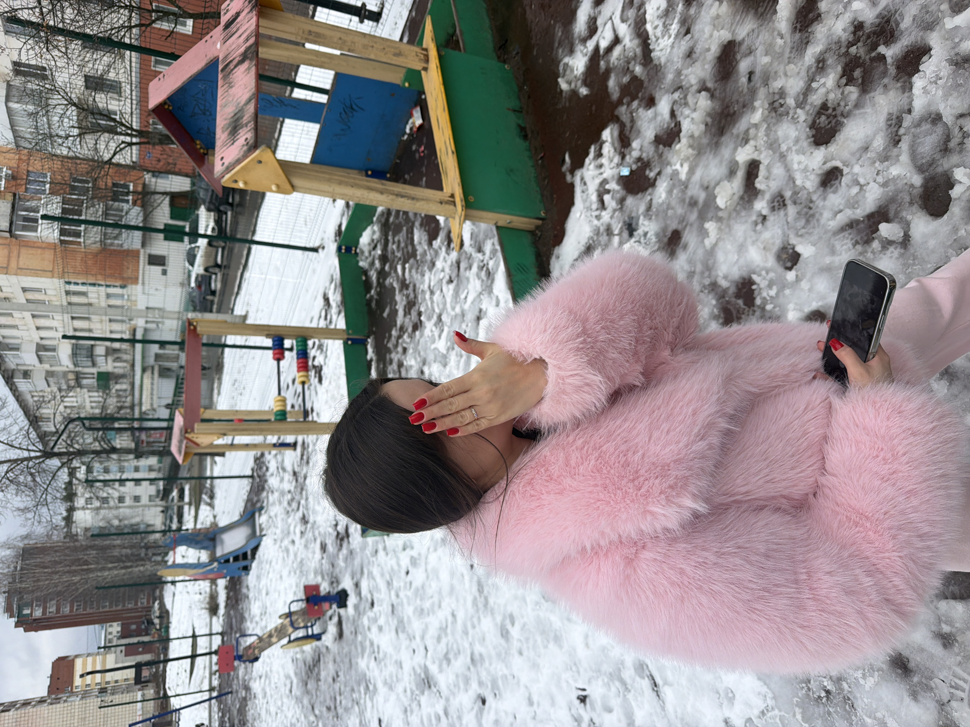 The image depicts a woman standing in a playground in Chaikovsky, Russia. The ground is covered with a mixture of snow and bare earth. She is wearing a pink fur coat and holding a smartphone. Her left hand covers part of her face, revealing red nail polish. Playground equipment is visible, including a small wooden structure with blue, yellow, and green elements, a slide, and a seesaw. Buildings are in the background, suggesting an urban environment.