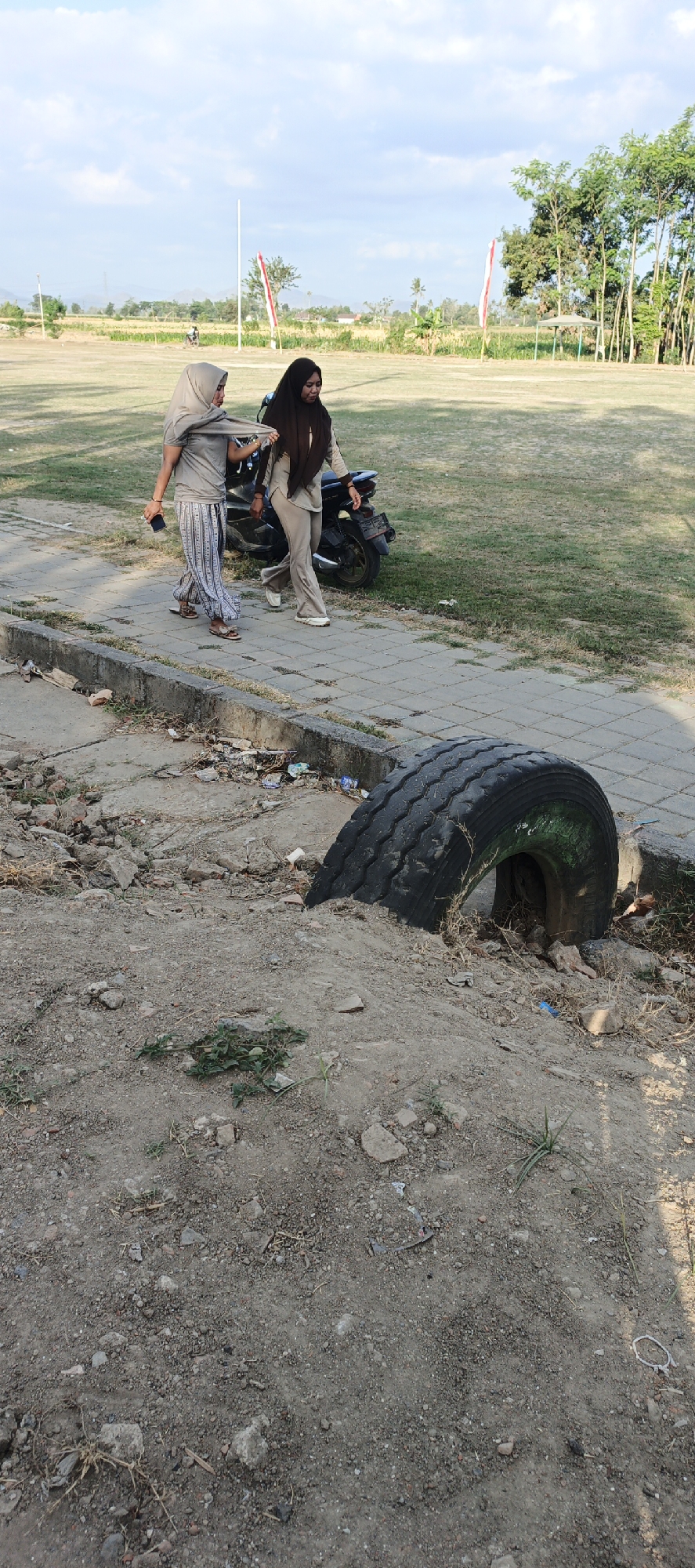 In Kasiyan Timur, Indonesia, a motorcycle is parked next to a paved walkway. Two women wearing hijabs are walking along the walkway, with one woman holding the other's arm. The walkway is bordered by a grassy field on one side and a dirt embankment on the other. A large, worn tire is partially embedded in the dirt embankment, with debris scattered around it. The sky is partly cloudy, and trees are visible in the background.
