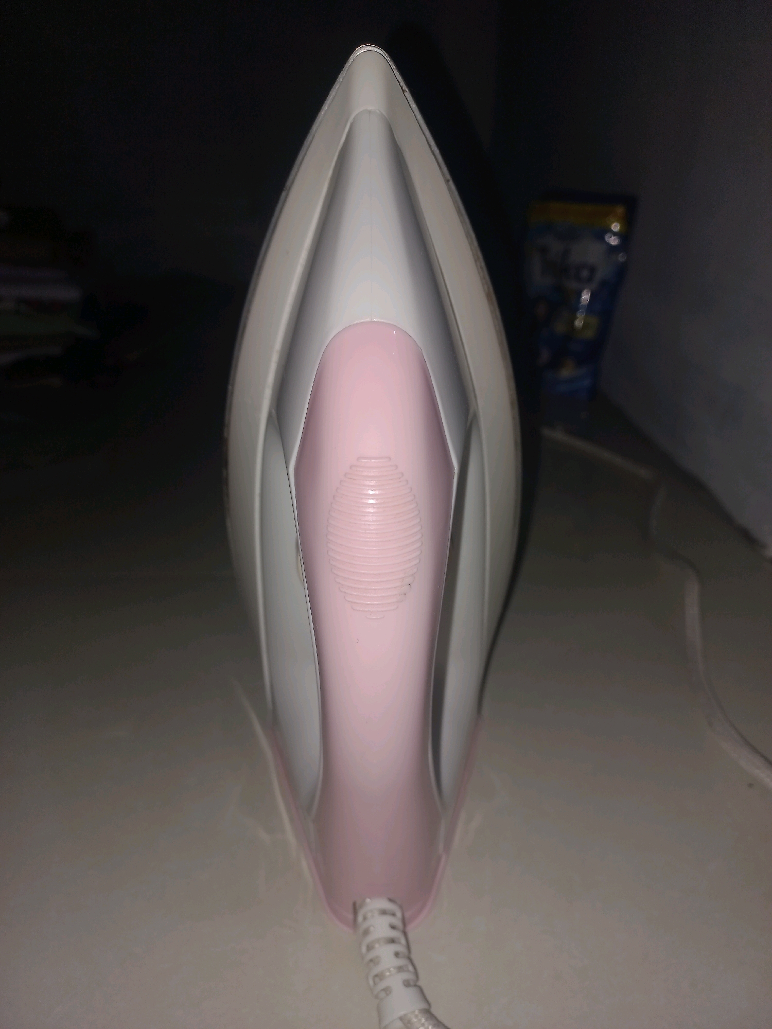 A close-up, top-down, eye-level shot focuses on the pointed front of a pink and white clothes iron. The iron is positioned vertically in the center of the frame, its tip facing upwards. The iron's body is a matte white with a distinct pink insert running down the center, which has a ridged texture. A white power cord, with a coiled section, extends from the bottom of the iron and disappears out of frame. The background is dark and out of focus, with a blurred blue and yellow pouch visible to the right of the iron, likely a cleaning product. The surface beneath the iron appears to be a light-colored, textured material, possibly a countertop. The lighting is dim, casting soft shadows. The image was taken in Gambiranom, Indonesia.