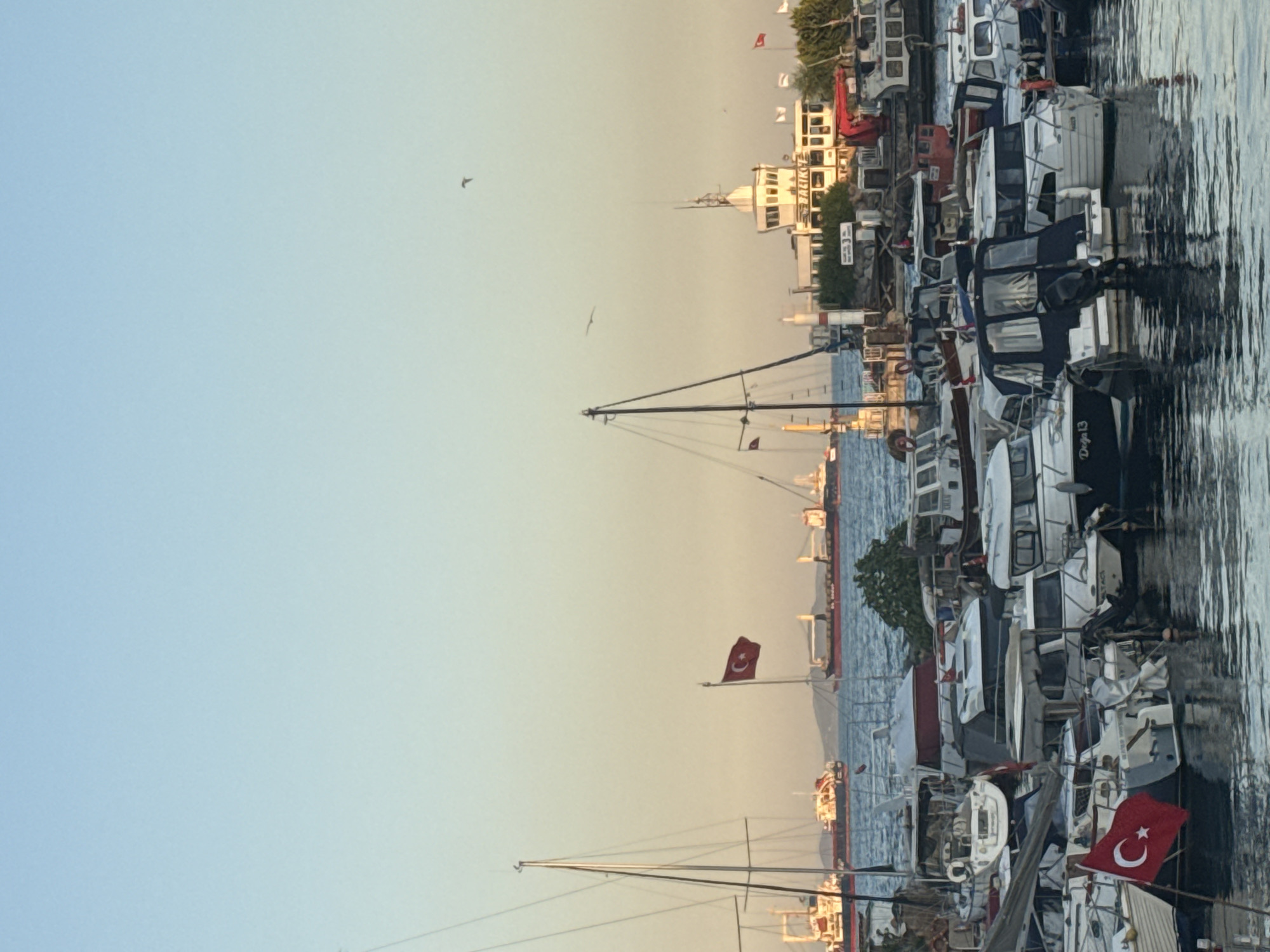 A long shot captures a harbor filled with numerous boats of various sizes. In the foreground, several small to medium-sized motorboats and sailboats are docked, some with their flags displayed. A prominent Turkish flag, red with a white star and crescent, is visible on one of the boats. In the background, larger cargo ships are anchored in the sea. A white tower building with a control room on top, likely a lighthouse or a maritime station, stands on the shore, surrounded by greenery. The sky is clear and transitions from a light blue at the top to a pale yellow-orange near the horizon, suggesting either sunrise or sunset. The water is calm, reflecting the boats and the sky. The scene is set in the city of Bakırköy, Turkey, as indicated by the text "BAKIRKOY" visible on the tower building. There are no visible people in the image.