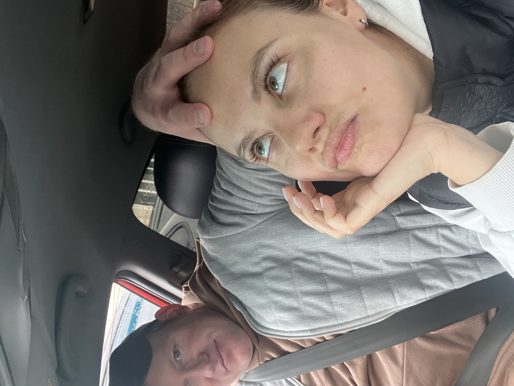 The image shows two people inside a car. A woman is in the foreground, partially reclined with her head supported by a hand on her forehead and another hand supporting her chin. She has light skin, light eyes, and light brown hair, wearing a light-colored top with a black vest. A man is visible in the lower portion of the frame, wearing a brown shirt and a black cap. Both individuals are wearing seatbelts. The interior of the car is visible, including parts of the dashboard and roof. A car wash is visible outside the car. It is not possible to determine the location in the city of Chaikovsky, Russia, from the given image.