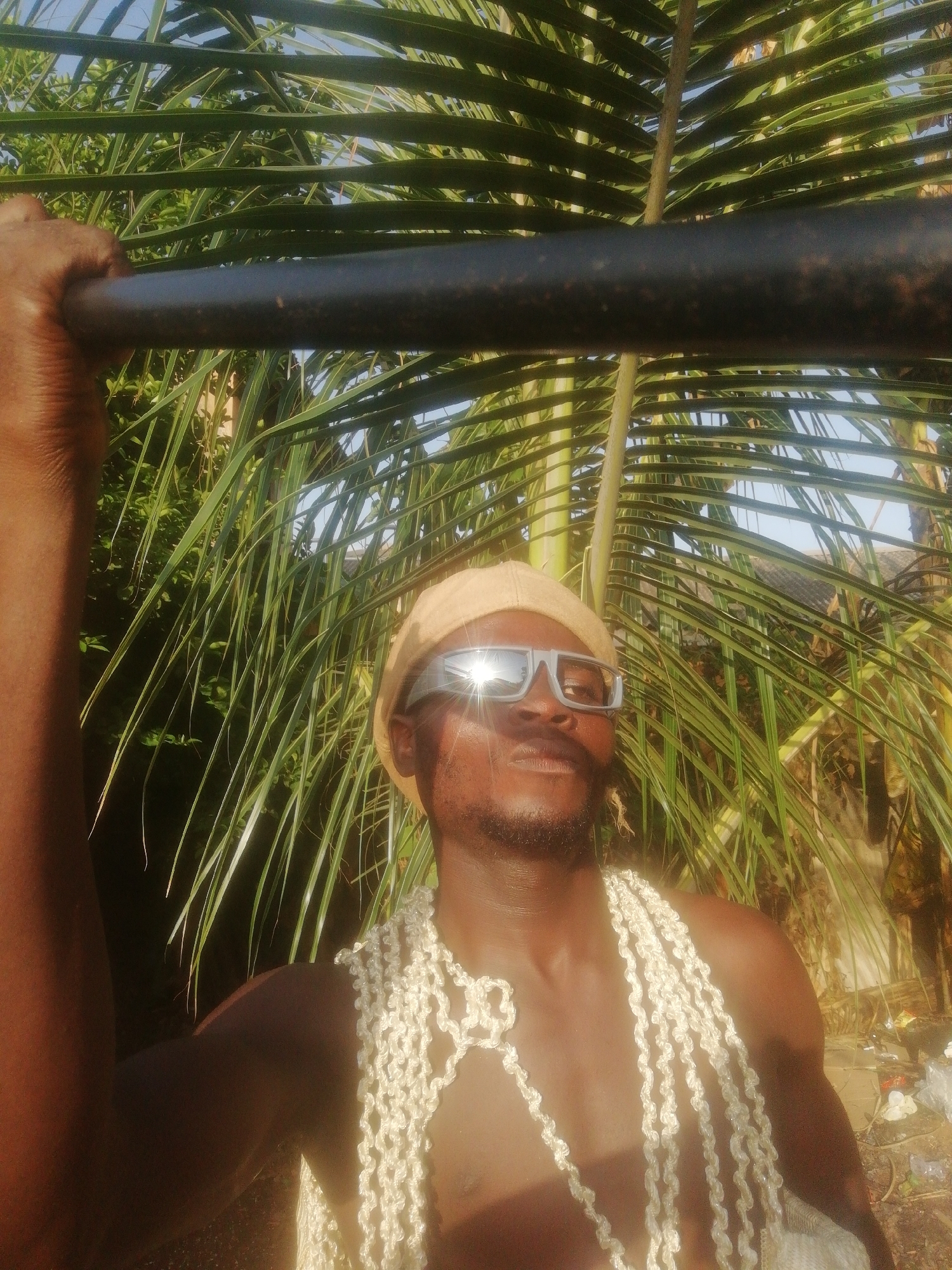 A dark-skinned man with facial hair and wearing mirrored sunglasses and a beige hat is seen from a low angle. He is also wearing what appears to be a garland of white shells or beads. He is holding onto a dark, round object, possibly a pipe or pole, with his left hand. The background consists of palm fronds and other foliage, suggesting a tropical or coastal location. The scene appears to be outdoors, likely in a natural environment.