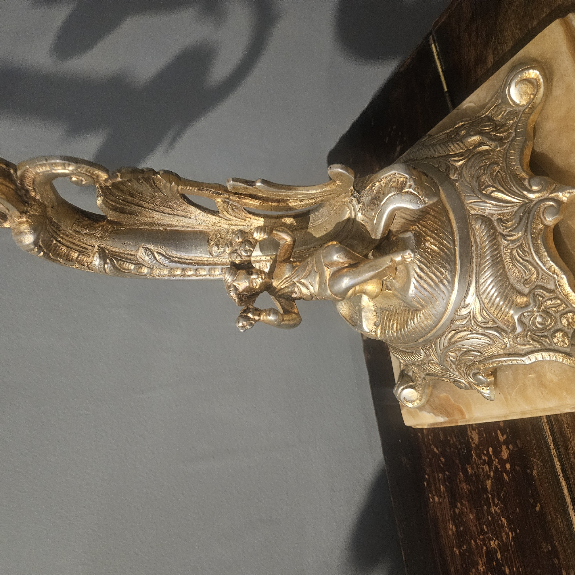 The image displays a close-up view of an ornate, antique metal figurine, possibly a candelabra base or decorative accent, resting on a polished wooden surface. The figurine is predominantly silver and gold in color, featuring intricate detailing. At the center, a small, seated cherubic figure with flowing drapery holds two clusters of what appear to be flowers or berries. The cherub is positioned on a decorative pedestal which itself sits on a polished, light-colored stone base, possibly marble or onyx. The metalwork of the figurine exhibits a distressed or antique patina, with some areas appearing darker or more worn than others, highlighting the sculpted textures and embellishments. The background consists of a plain, grey wall, against which shadows from an unseen light source cast elongated, organic shapes, suggesting the presence of foliage or abstract forms. The lighting is dramatic, creating strong contrasts between the illuminated parts of the figurine and the shadowed areas. The overall impression is one of historical craftsmanship and artistic display. No specific location in Almadrones, Spain, can be determined from the image itself, as it focuses solely on the object and its immediate surroundings.