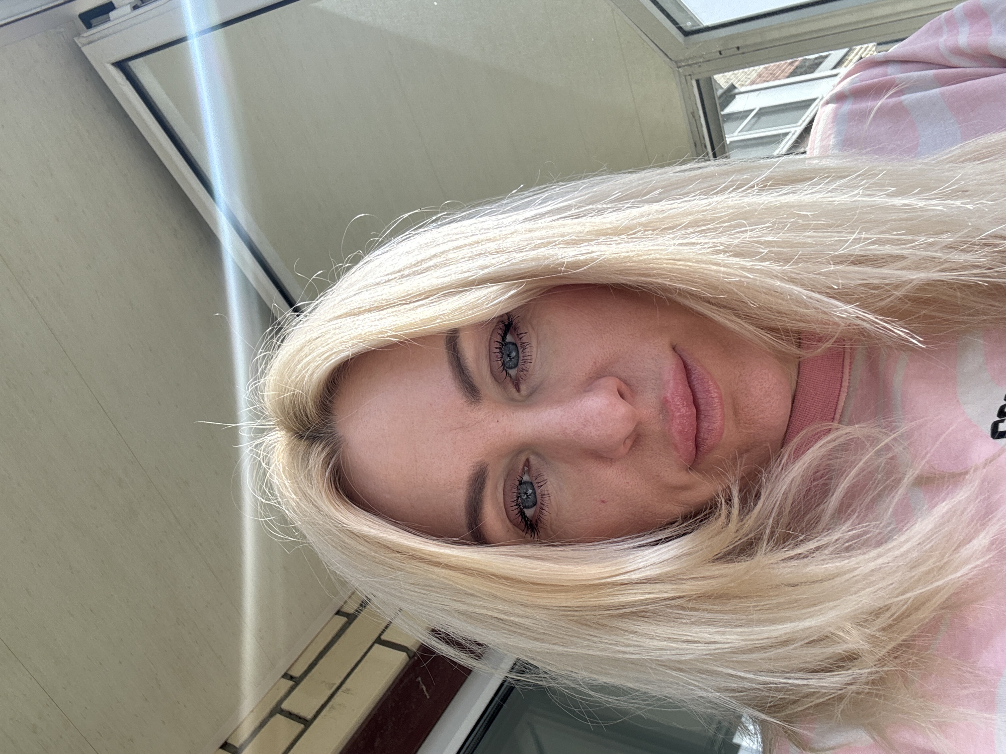 A close-up, canted angle shot focuses on a woman with blonde hair and blue eyes. The woman is wearing a pink and white striped top. The background consists of a building exterior and a window, which suggests an indoor setting. The location is listed as the city of Unknown, Unknown.