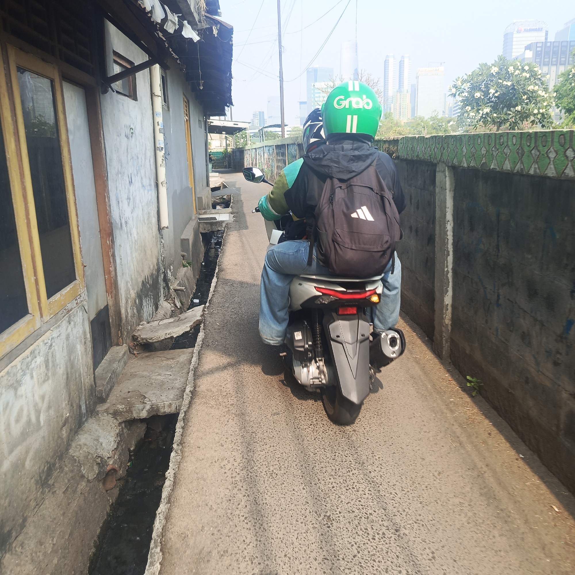 Two individuals are riding a scooter down a narrow, paved alleyway. The person in the back is wearing a green Grab helmet, indicating they are likely a ride-hailing service driver. Both individuals are wearing backpacks. The alley is flanked by weathered, gray buildings on the left and a textured concrete wall on the right. A drainage ditch runs alongside the buildings, filled with dark, murky water. In the background, modern skyscrapers are visible, suggesting an urban environment in the distance. The lighting indicates it is daytime, with sunlight casting shadows on the ground. The overall scene depicts everyday life in a densely populated urban area.