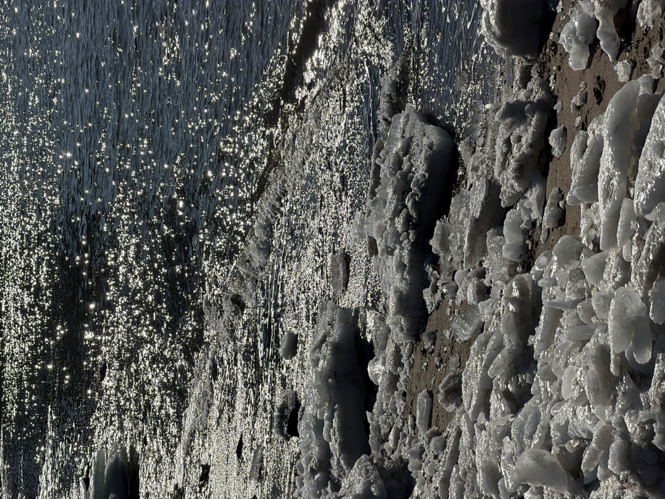Sunlight dances on a choppy body of water, creating glittering reflections across its surface. Along the shore, a mixture of ice and sand creates a textured landscape. Large formations of ice cling to the beach, sculpted by the cold and the constant push of the waves. The ice glistens in the light, juxtaposed against the dark and shimmering water.