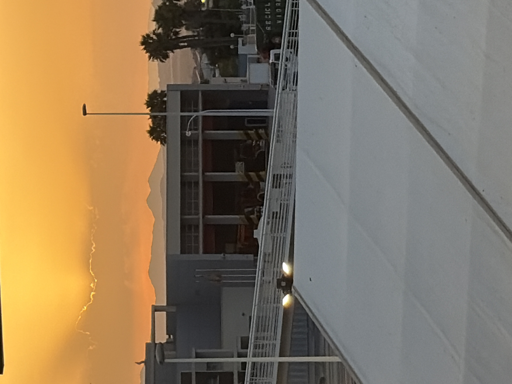 The image depicts a sunset over Torrevieja, Spain. The sky is a vibrant orange, with the sun partially obscured by clouds, creating a dramatic effect. In the foreground, a white tent-like structure occupies the bottom portion of the frame. Behind this, a modern building complex is visible, featuring grey and white facades. A ramp or walkway with white railings leads up to the building. Streetlights are present on the walkway, with two emitting a bright yellow light. Further in the background, palm trees and distant mountains are silhouetted against the colorful sky. A green dumpster with the label "RECICLAVIL" is also visible. The scene appears to be an outdoor area, possibly a port or industrial zone, given the presence of large vehicles and structures.