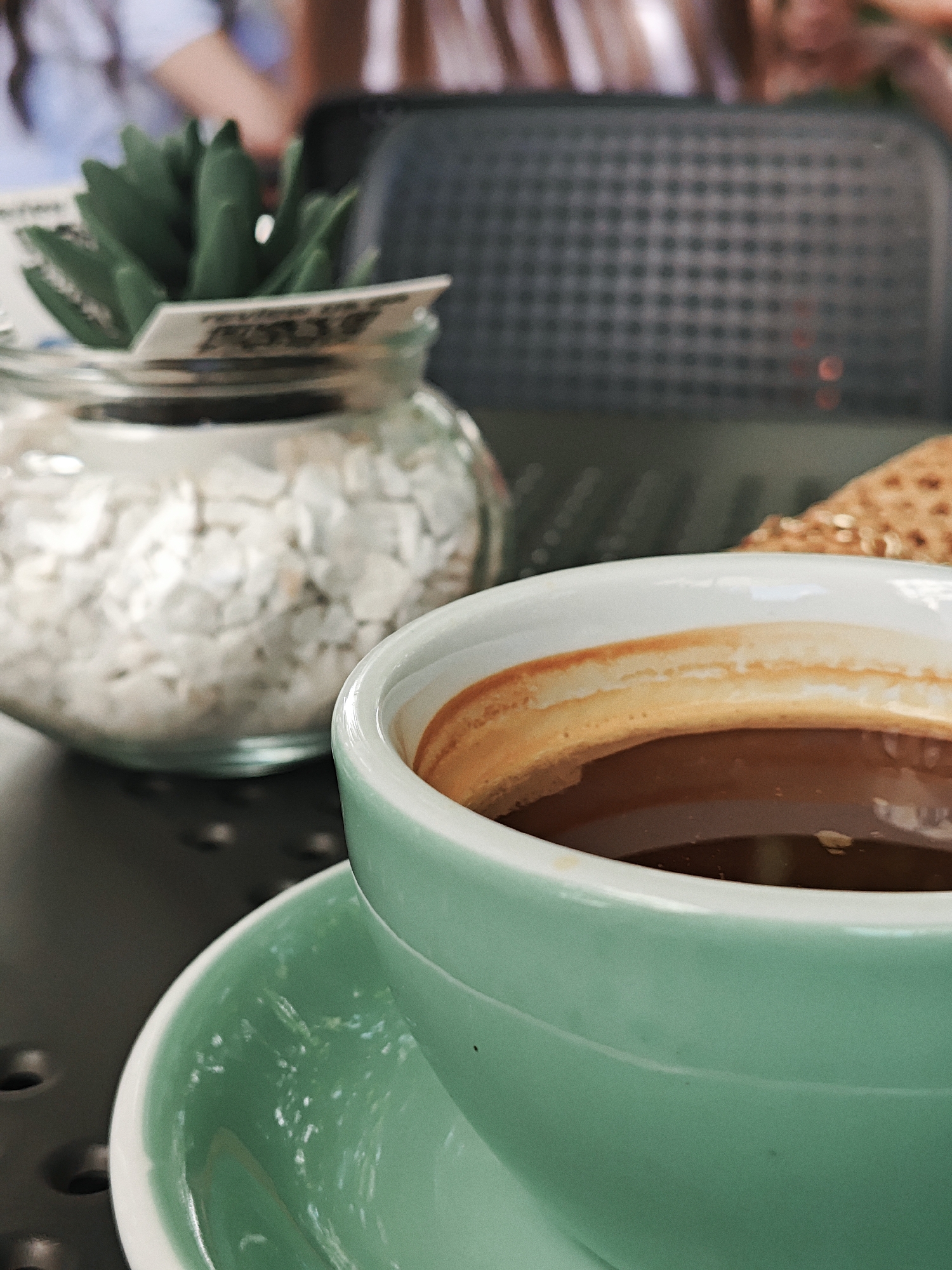 The image shows a close-up of a teal coffee cup and saucer filled with dark coffee, set on a dark, perforated surface. To the left of the coffee cup, partially out of focus, is a glass jar containing white pebbles and a small artificial succulent plant. In the background, also out of focus, is a black, textured object, possibly a tray or a grill. The overall scene suggests a casual cafe setting.