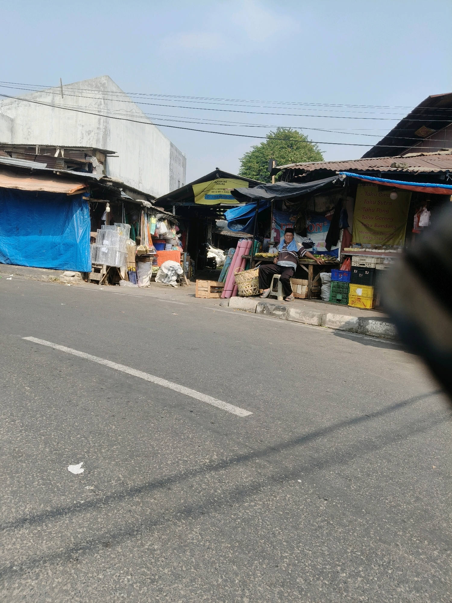 The image depicts a street scene in South Tangerang, Indonesia, likely a commercial area with roadside stalls. In the foreground, a grey asphalt road with a white dividing line is visible. The midground features a row of market stalls constructed from corrugated metal and tarps. One stall has a bright blue tarp covering it, and another displays white wire racks. A man, dressed in a striped polo shirt and dark pants, sits on a stool in front of a stall, appearing to be a vendor. He is leaning on a wooden table laden with produce, including bananas. Several plastic crates, some stacked and colored yellow, green, and blue, are placed on the sidewalk. Behind the stalls, a large, white, angular building is visible, contrasting with the rustic market structures. A lush green tree rises behind the stalls, suggesting a somewhat urban environment with pockets of vegetation. The sky is clear and blue, indicating a sunny day. The overall impression is of a busy, informal marketplace operating alongside a public road.