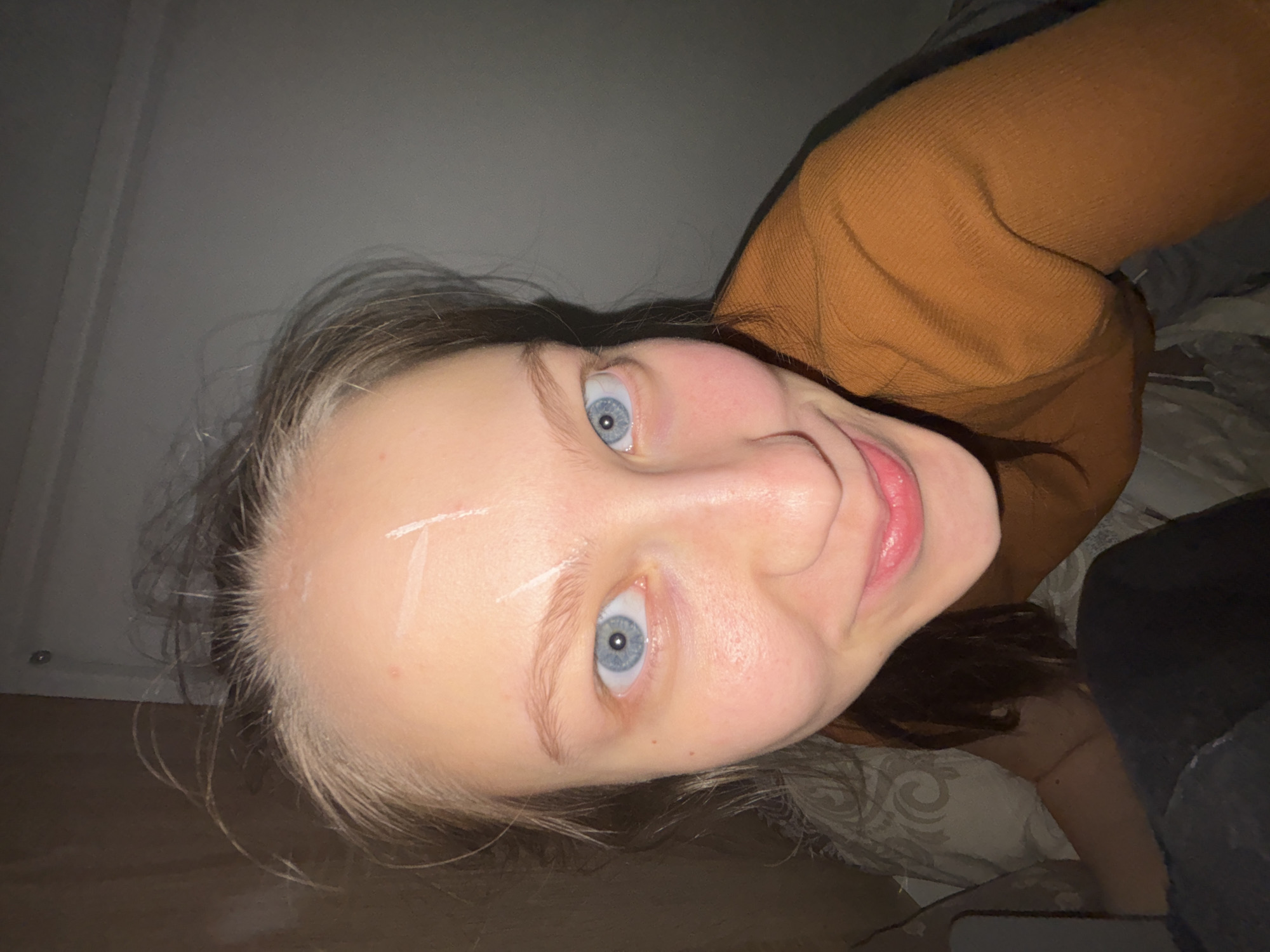 A person is lying down, oriented with their head towards the upper left corner and their body extending diagonally towards the lower right. The person has fair skin, light blonde hair with darker roots, and light blue eyes. There appears to be a small wound covered with adhesive bandage strips on the forehead. They are wearing a brown, knit sweater. The person is lying on a bed with a patterned, light gray duvet. The background is indistinct and appears to be a dimly lit room. The location is not discernible from the image.