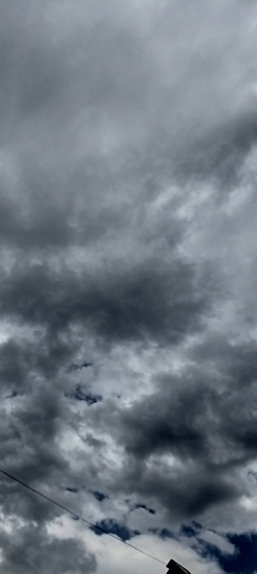 The image shows a sky filled with dark, ominous storm clouds. A thin wire runs diagonally across the lower part of the frame, disappearing into the bottom right corner where the edge of a dark, angled rooftop is visible. The overall scene suggests an impending storm in the city of Sokuluk, Kyrgyzstan, characterized by the dramatic cloud formations and a hint of human infrastructure.