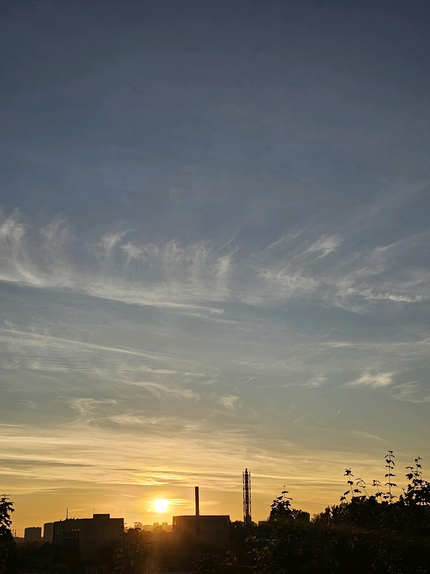 The image captures a sunset over the city of Khimki, Russia. The sun is a bright, orange orb low on the horizon, casting a warm glow across the sky. Wispy, white clouds streak across the upper half of the frame, contrasting with the deepening blue of the sky. In the foreground, silhouetted against the vibrant sky, are the rooftops of buildings, a tall industrial smokestack, and the dark shapes of trees. The overall scene depicts an urban landscape bathed in the soft light of dusk.