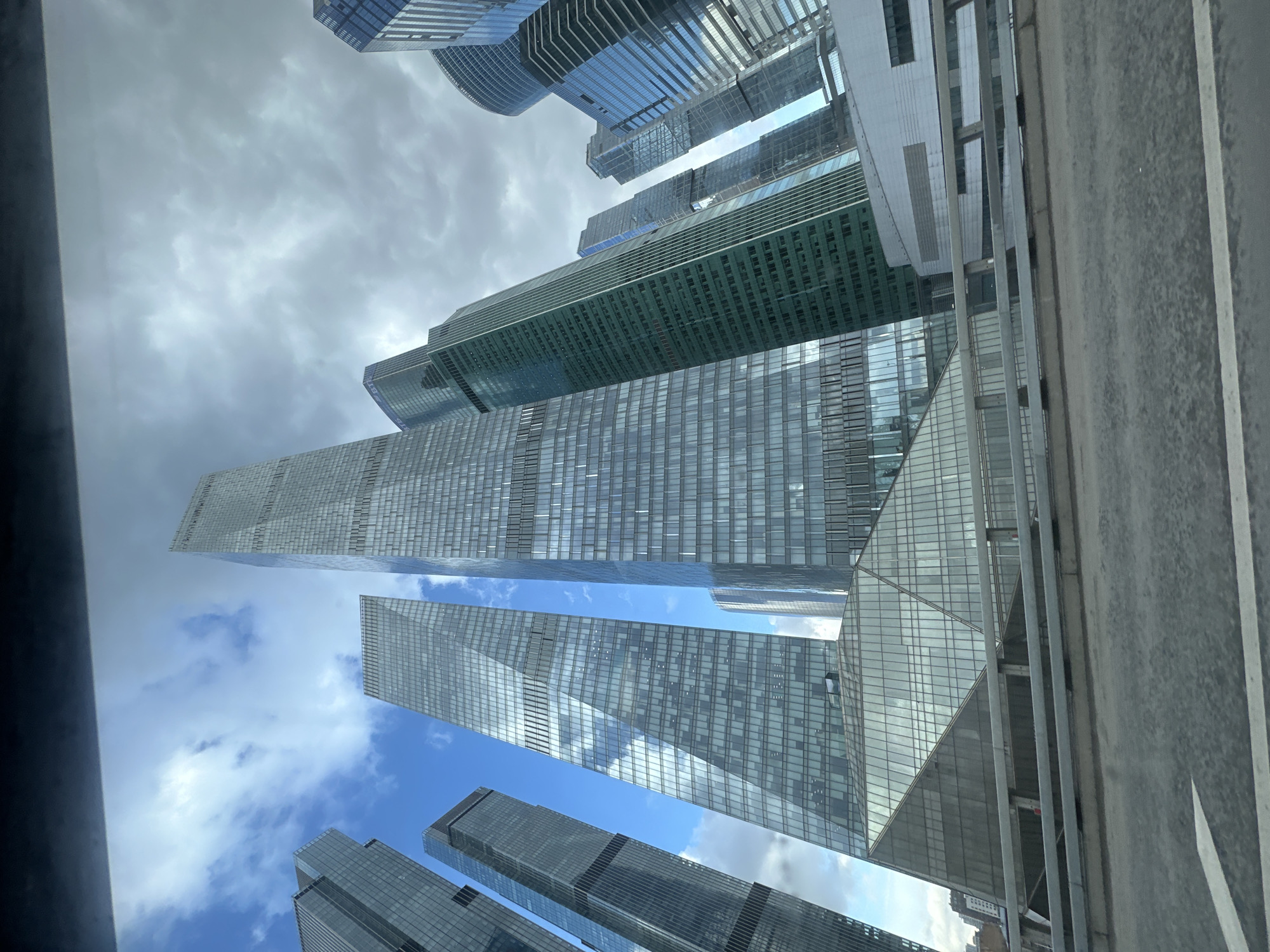 The image shows a low-angle view of the Moscow International Business Center (Moscow-City) in Moscow, Russia. Tall skyscrapers with glass facades dominate the scene, reflecting the sky and clouds. The sky appears partly cloudy with patches of blue. The angle suggests the photograph was taken from ground level, looking upward. Some structural elements, possibly from a bridge or elevated roadway, are visible at the right of the image, indicating the photographer's position. No people are visible.