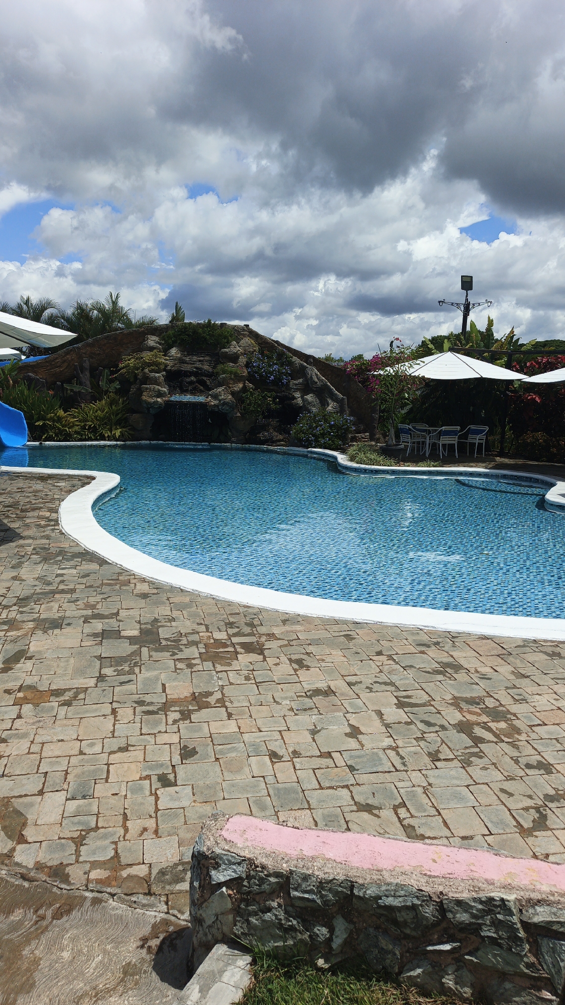 This image presents a serene outdoor swimming pool area, likely part of a resort or leisure facility, under a partly cloudy sky. The central feature is an irregularly shaped swimming pool filled with clear, blue water, tiled with small mosaic patterns. A bright white coping outlines the pool's edge, separating it from the surrounding paved area.
To the left, a vibrant blue water slide curves downwards, suggesting aquatic recreational opportunities. Behind the pool, a natural-looking rock formation features a small waterfall cascading into the water. This rockery is adorned with diverse green foliage, including bushes, plants, and palm trees, along with scattered colorful flowers in shades of purple and pink/red, enhancing a tropical aesthetic.
On the right side of the pool, a shaded seating area is visible, complete with a white outdoor umbrella, a table, and chairs, indicative of a place for relaxation. A tall lamppost or light fixture stands in the background on this side.
The extensive patio surrounding the pool is paved with irregularly shaped stone pavers, displaying a mix of grey, brown, and reddish hues. Some wet patches on the paving suggest recent water activity or a damp environment. In the immediate foreground, a low, rugged stone wall or bench is present, topped with a distinctive pink painted surface, with a small patch of green grass at its base.
The sky is characterized by a mix of large, fluffy grey and white clouds, with intermittent patches of clear blue sky visible, indicating a bright but partly overcast day. The absence of people in the scene contributes to a tranquil and inviting atmosphere. The overall setting implies a destination for leisure and enjoyment, located in Vallecito, Venezuela. No visible text is present in the image.