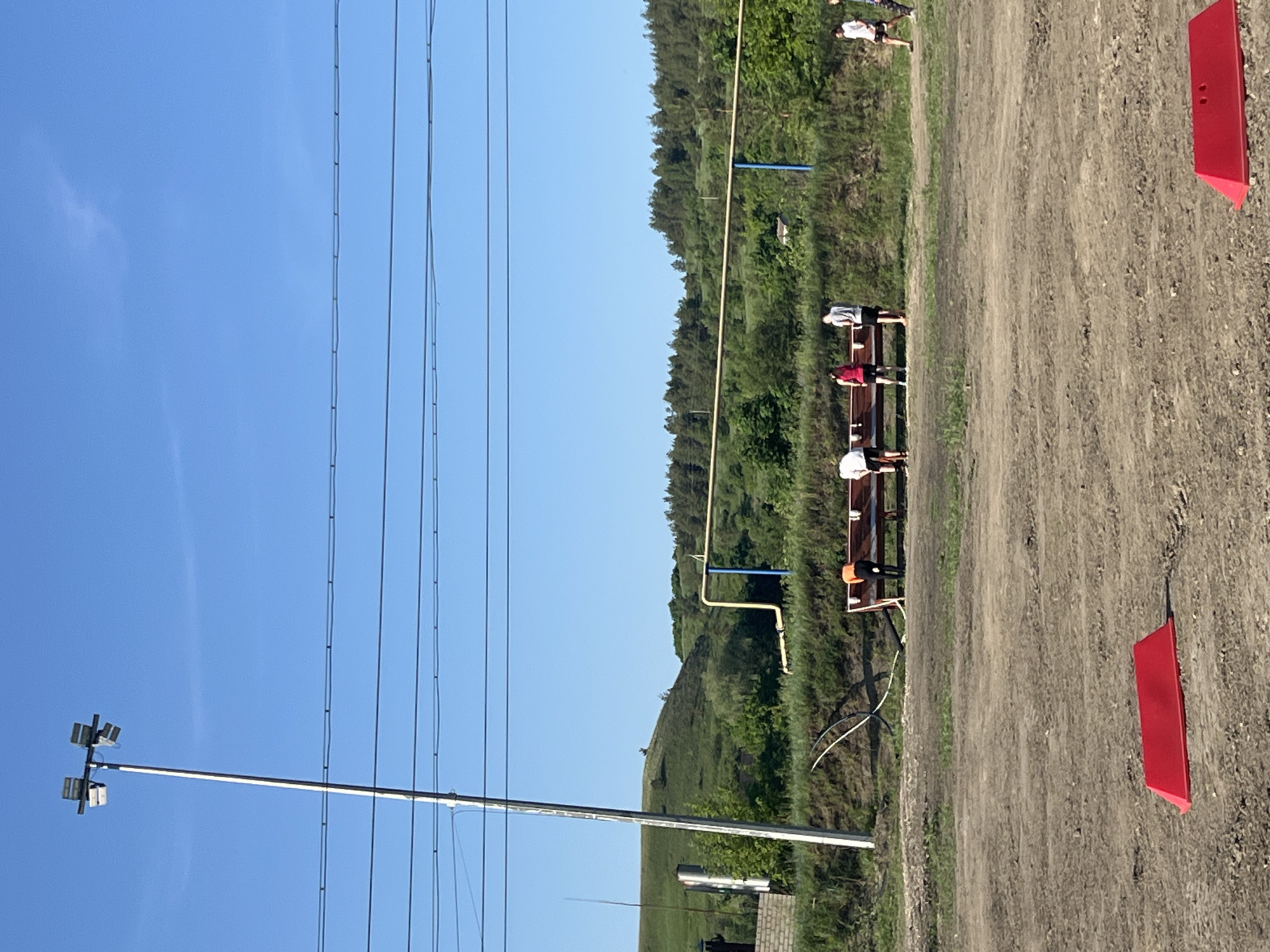The image depicts an outdoor scene in Khvalynsk, Russia. The environment includes a clear blue sky, grassy terrain, and a dirt track. Overhead power lines are visible. A group of people stands near a wooden structure, possibly a bench or platform. Red rectangular markers are placed along the dirt track. In the background, a hill or embankment is covered with trees. A utility pole with lights is also present.