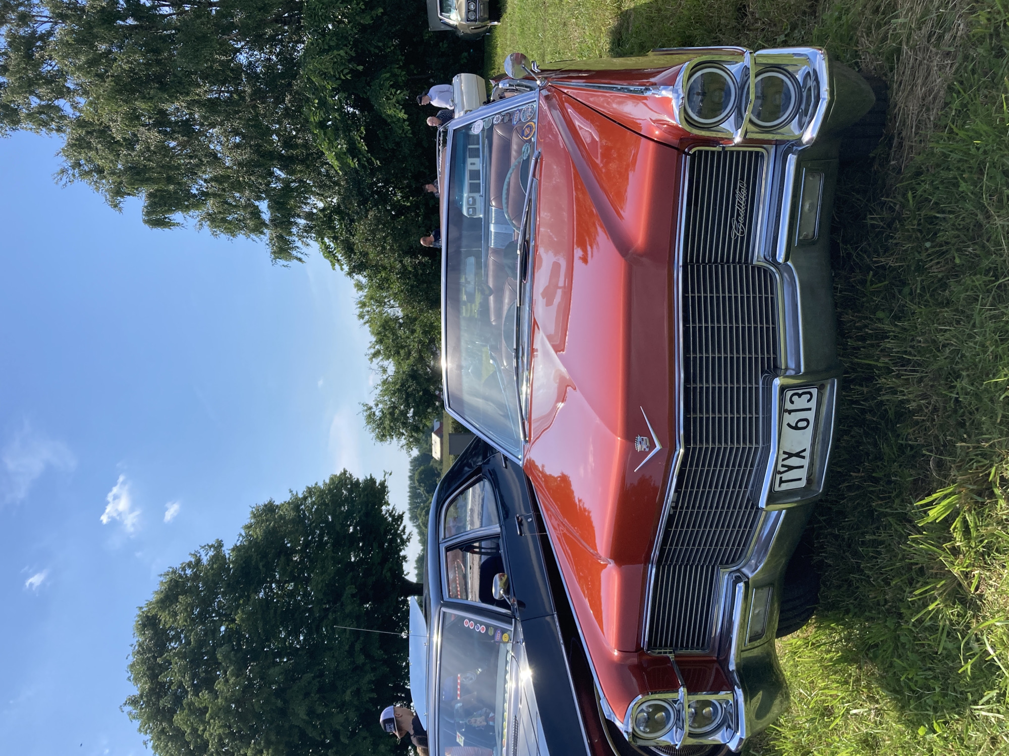 The image shows a classic red Cadillac convertible parked on a grassy area. The car has chrome accents and a prominent grill. Behind the Cadillac, a black sedan is visible, and further back, several people are standing near other vehicles and trees. The sky is blue with scattered clouds. The scene appears to be an outdoor event or car show, possibly in Edenberga, Sweden, given the context.