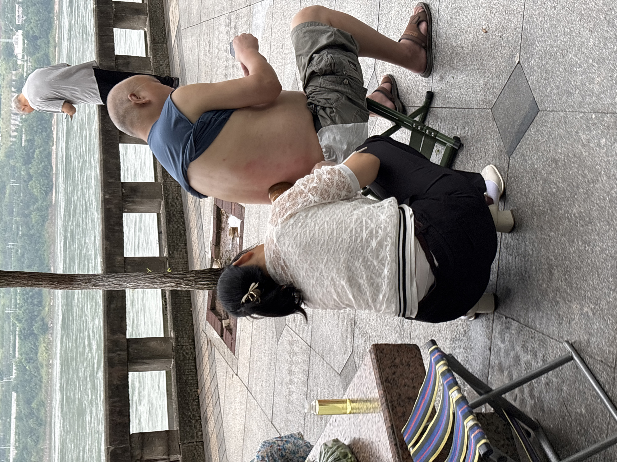 The image depicts a scene on a paved walkway along a body of water, likely the Yangtze River in Jiangjin, China. In the foreground, a woman is kneeling and using a wooden implement to massage the back of a man who is sitting on a small green folding chair. The man's back is exposed, revealing red marks, possibly from the massage. He is wearing a blue t-shirt and khaki shorts, and sandals. The woman is wearing a white lacy top and black pants, with white socks and light-colored heeled shoes. To the left, there is a small folding chair with a striped fabric seat and a granite-topped table with a small bottle of yellow liquid and a green plastic bag. In the background, another man in a light grey t-shirt and dark pants stands near a stone railing, looking at his phone. Beyond the railing is a wide river, and further in the distance, a green, hilly landscape. The overall atmosphere suggests a public space where relaxation and personal care are taking place.