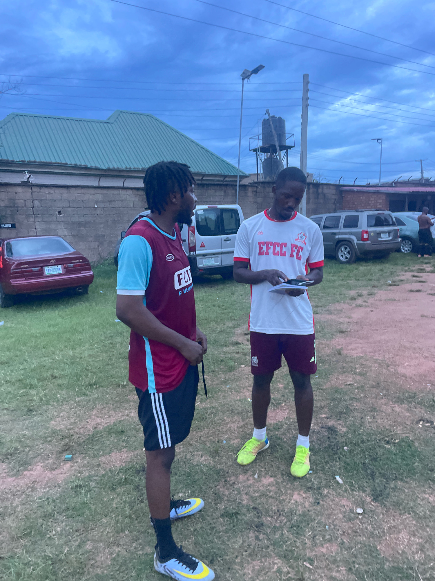 Two young Black men are standing on a grassy field under an overcast sky, likely in the city of Jos, Nigeria. The man on the left wears a maroon and light blue football jersey with white trim, black shorts with three white stripes, black socks, and light blue and yellow football cleats. He has dreadlocks. The man on the right wears a white football jersey with red trim and the text "EFCC FC" in red, maroon shorts, and bright yellow football cleats. He is looking down at a tablet or notebook he is holding. In the background, there are several parked vehicles, including a red sedan and a gray SUV, a brick wall, a building with a green roof, a water tower, and a lamppost. The overall lighting suggests it is late afternoon or early evening.