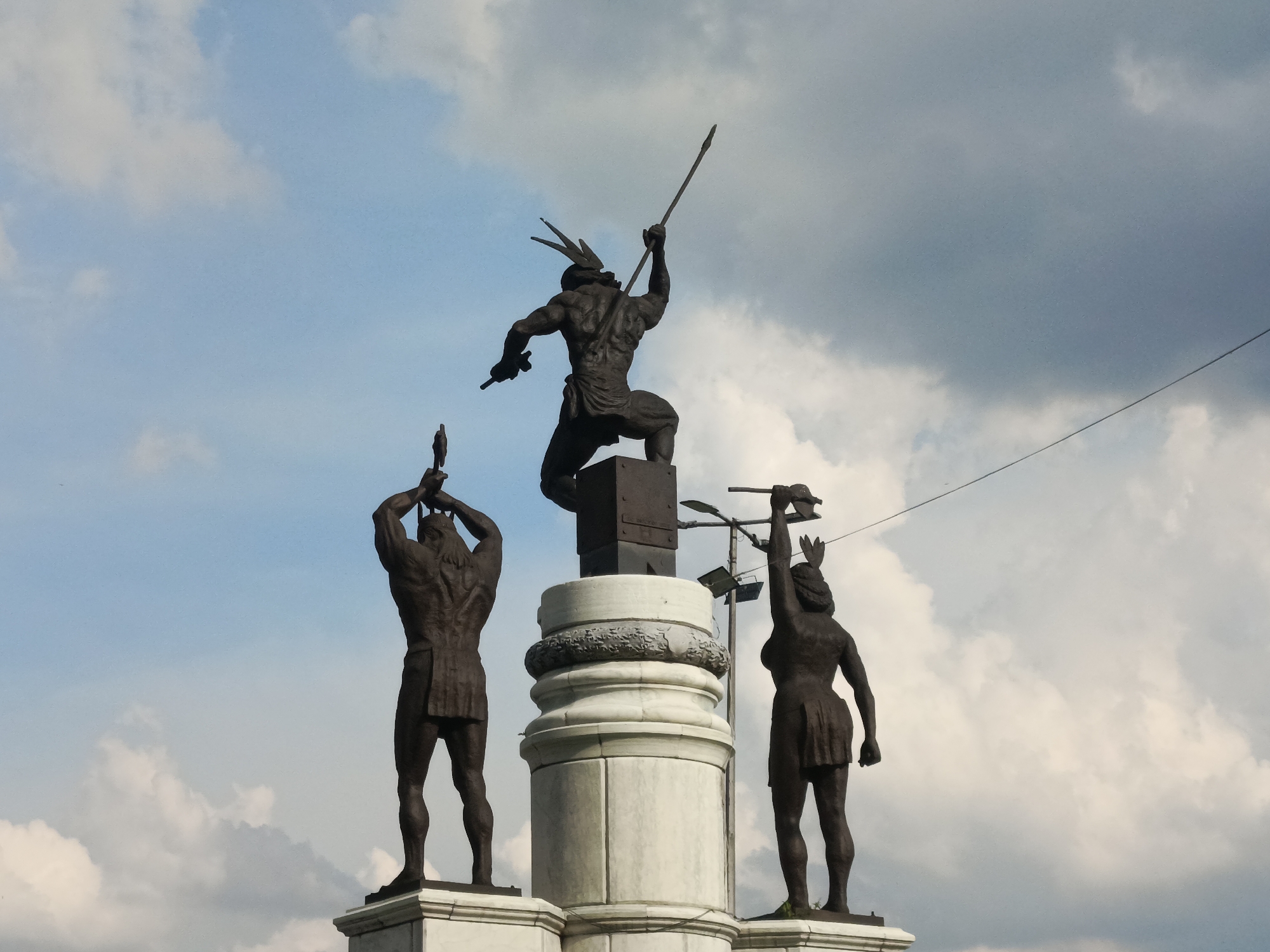The image shows the indigenous resistance monument erected in Caracas, Venezuela. Three bronze statues of naked indigenous men stand on pedestals surrounding a larger one. Each figure brandishes a spear. The sky is cloudy. A power line is visible.
