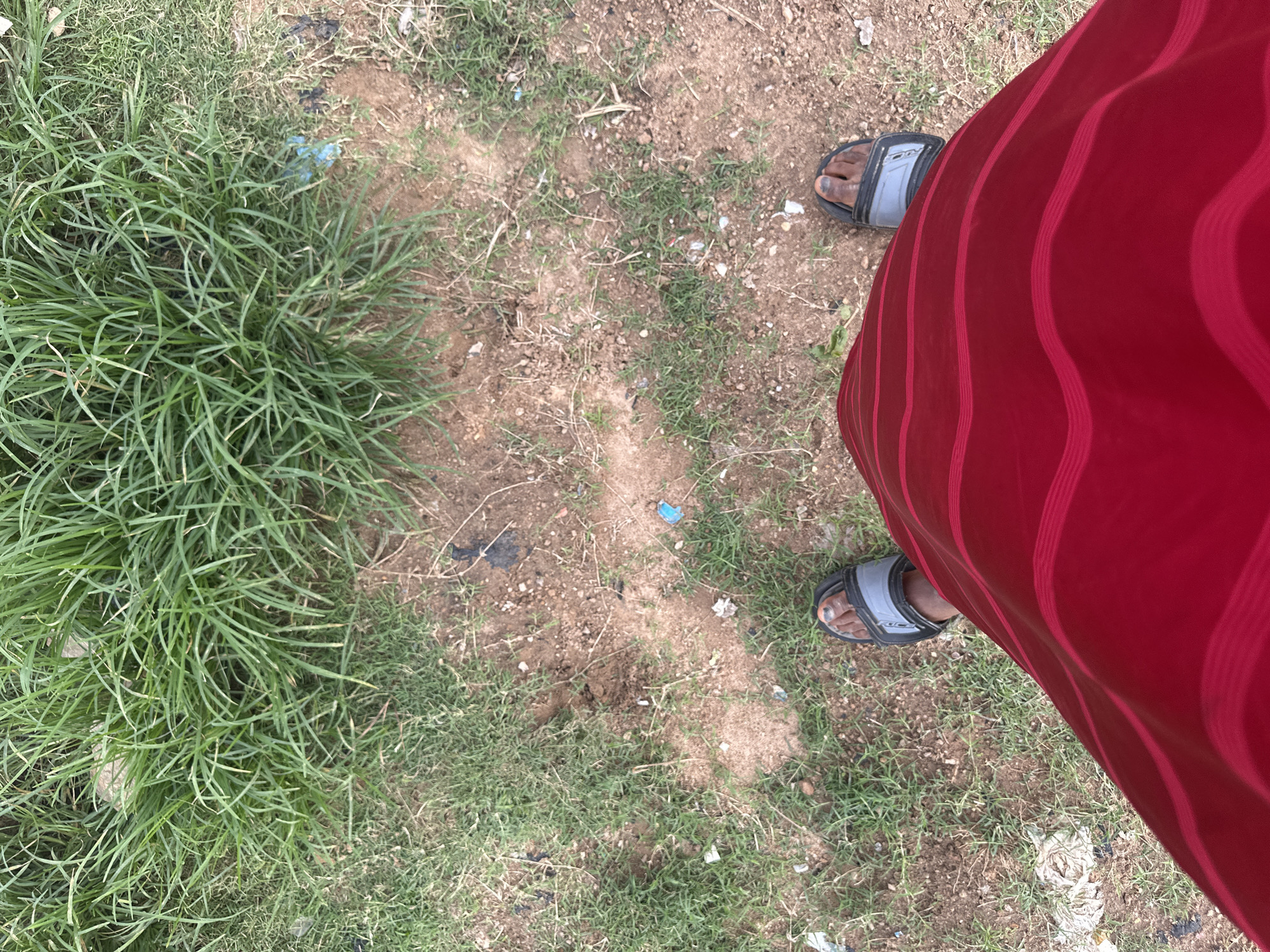 A ground-level, eye-level shot captures a person's legs and feet standing on a patch of dirt and grass on Rukuba Road, Nigeria. The individual is wearing a red skirt with horizontal pink stripes and black and grey sandals. Their feet are positioned on a mix of dry soil, sparse green grass, and a few scattered pieces of litter, including a small blue plastic item. In the upper portion of the image, a denser growth of vibrant green grass covers the ground. The overall scene is outdoors, with evidence of a somewhat unkempt environment.