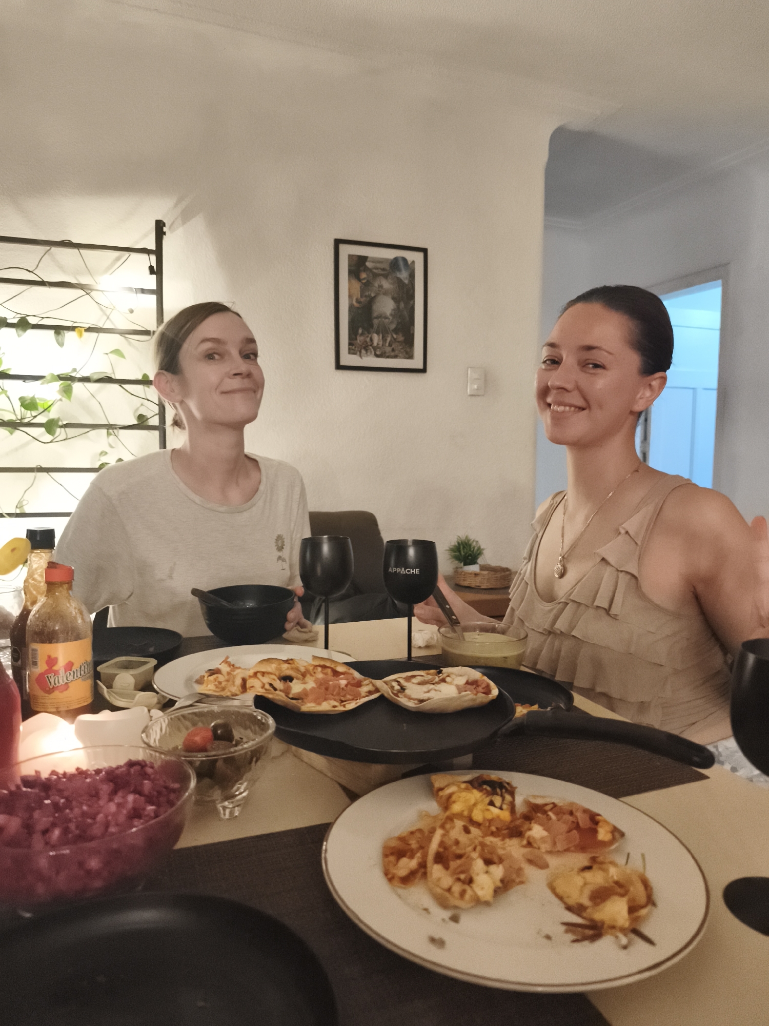 Two women are seated at a table indoors. The woman on the left wears a light-colored t-shirt, while the woman on the right wears a sleeveless, ruffled beige top and a gold necklace. They are both smiling. On the table are plates of food, including what appears to be pizza, a bowl of red chopped food, bottles of hot sauce, and glasses with the word "Appache" on them. There is a small potted plant on a shelf in the background. A painting hangs on the wall. The photo was taken indoors in Mexico City, Mexico.