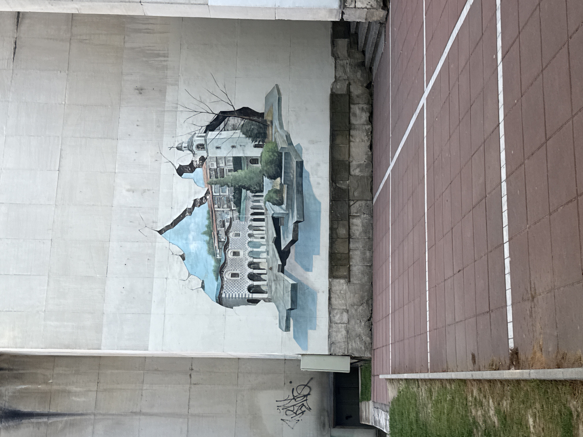 The image shows a mural on a concrete structure, likely a bridge support, adjacent to a paved walkway. The mural depicts a building or complex of buildings in a style that appears to break through the concrete surface. The artwork includes architectural details, foliage, and a blue sky. There is graffiti on the concrete below the mural. The walkway is made of brown pavers with white dividing lines. Grass and some kind of structure are visible at the edge of the walkway. The mural is located at the pedestrian tunnel in front of the American University in Blagoevgrad, Bulgaria.