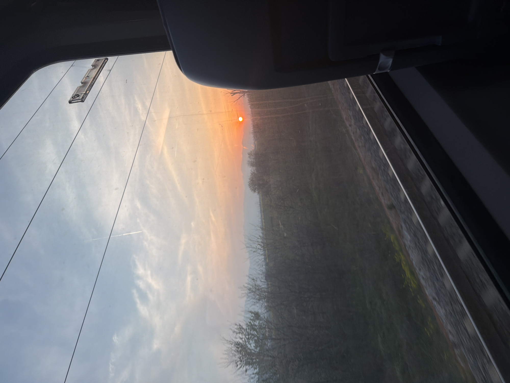 The image, taken from inside a vehicle, shows a landscape at sunset or sunrise through a window. The sky is a blend of blue and orange hues, transitioning from a lighter blue with clouds to a warm orange where the sun is visible. Below the sky, a line of trees and foliage is visible, with some details obscured by the window's transparency and potential moisture. The window frame and interior components of the vehicle are present along the edges of the frame, indicating the perspective of the shot. The scene appears to be a natural setting, possibly on the outskirts of a city.
Without additional information, it is impossible to definitively place this scene within the city of Plovdiv, Bulgaria. There are no identifiable landmarks, architectural features, or specific geographical details to conclusively confirm this location.
