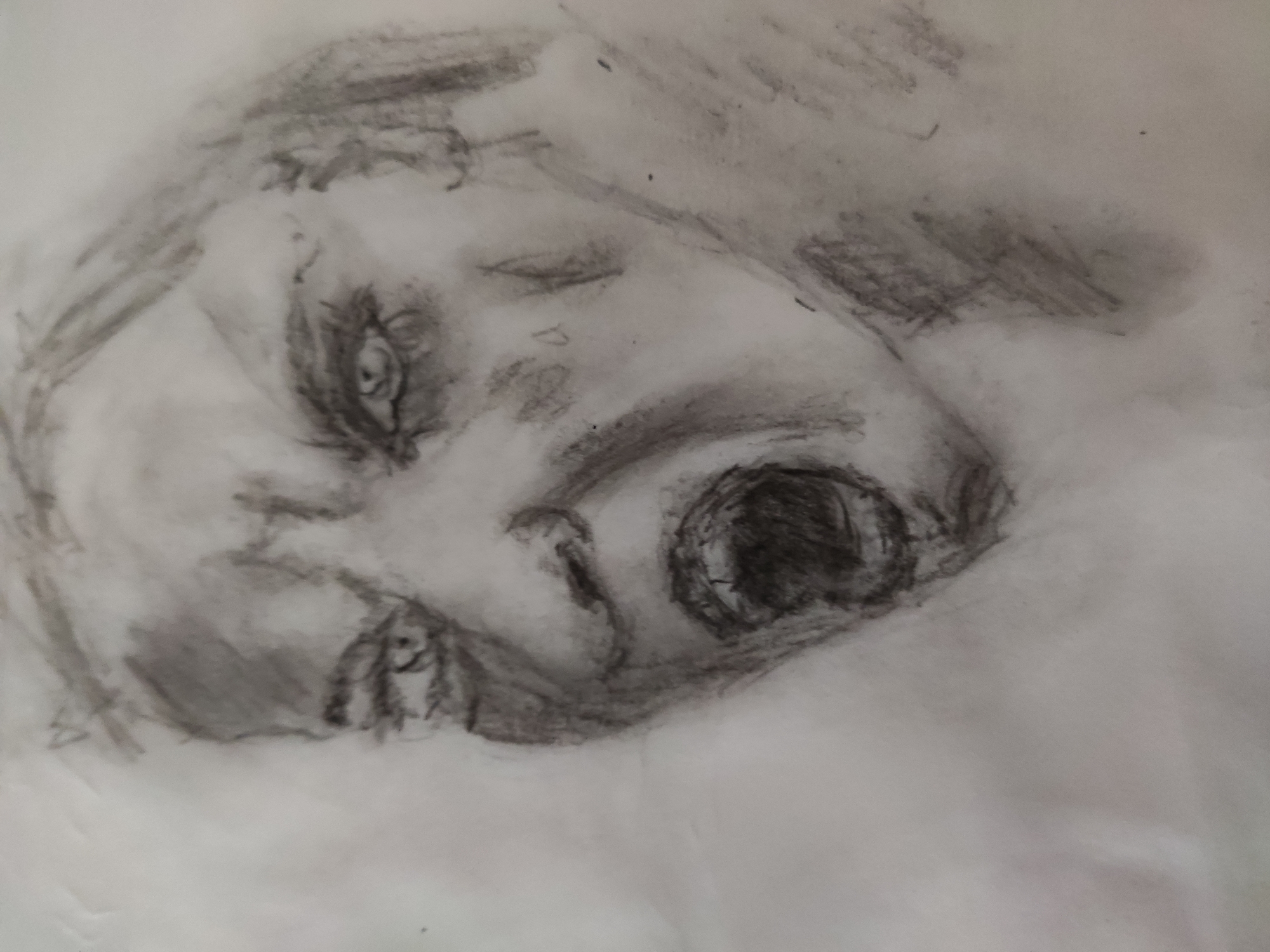 A charcoal drawing on paper depicts a person's face in close-up, with their mouth wide open in a scream or shout. The drawing is rendered in shades of gray, with bold lines and smudging creating a sense of raw emotion and intensity. The eyes are wide and focused, conveying a sense of distress or anger. The hair is loosely sketched, framing the face. The background is largely undefined, with a gradient of lighter to darker gray, drawing full attention to the expressive face. The drawing appears to be unfinished or in a sketch phase, with visible pencil marks and areas of lighter shading. The artwork is not located in a specific city or environment, as it is a drawing on paper.