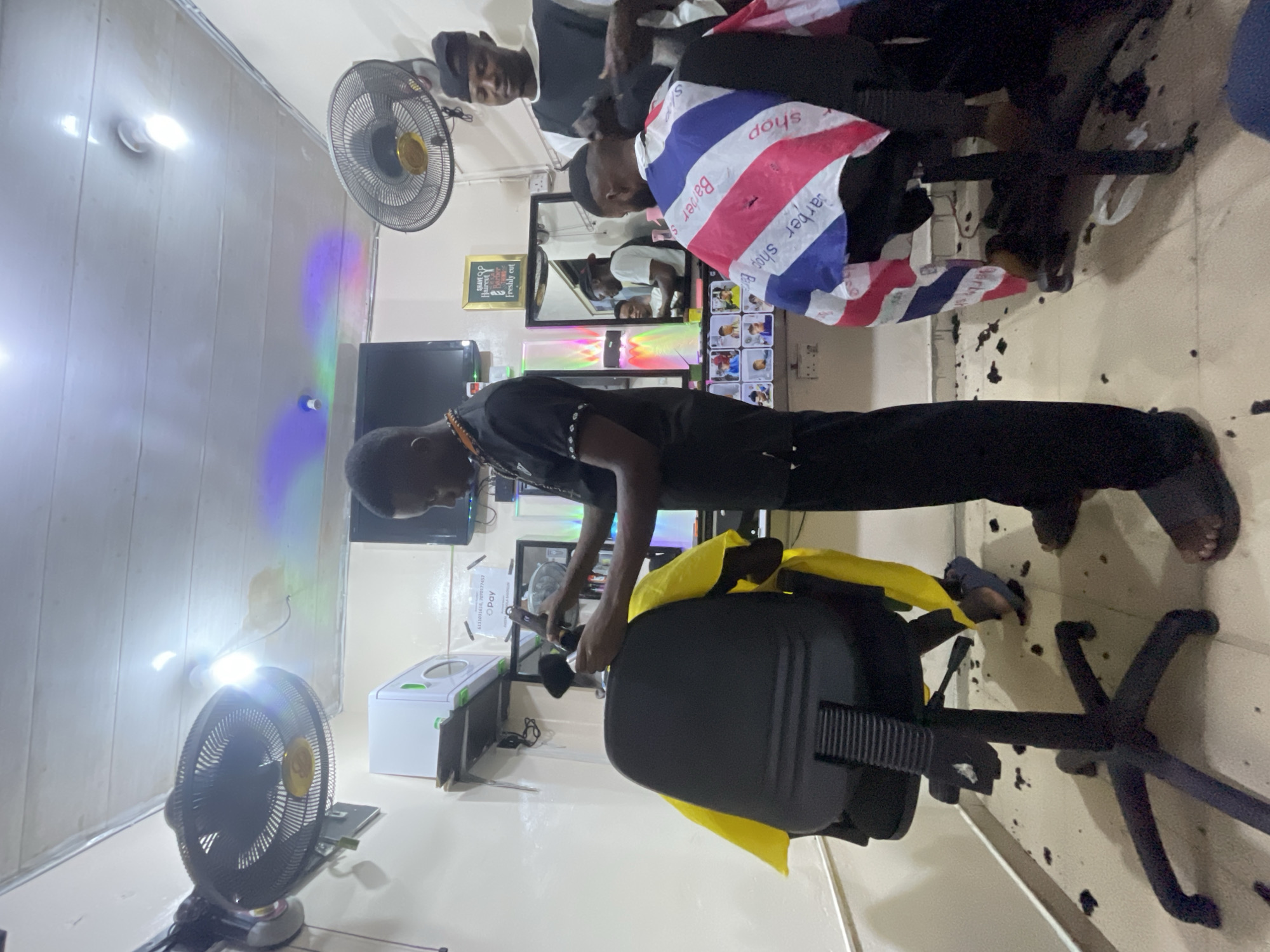 The image shows the interior of a barbershop in Ado, Nigeria. A barber dressed in black is giving a haircut to a seated customer. Another barber is attending to a customer in the background. A blue, white, and red cape is draped around the neck of the customer in the background. Hair clippings are scattered on the floor. There are two standing fans in the shop, one near the front and one near the back. Mirrors and posters are visible on the walls, along with illuminated signage and a television. A hygiene cabinet is located along the wall, above the floor.