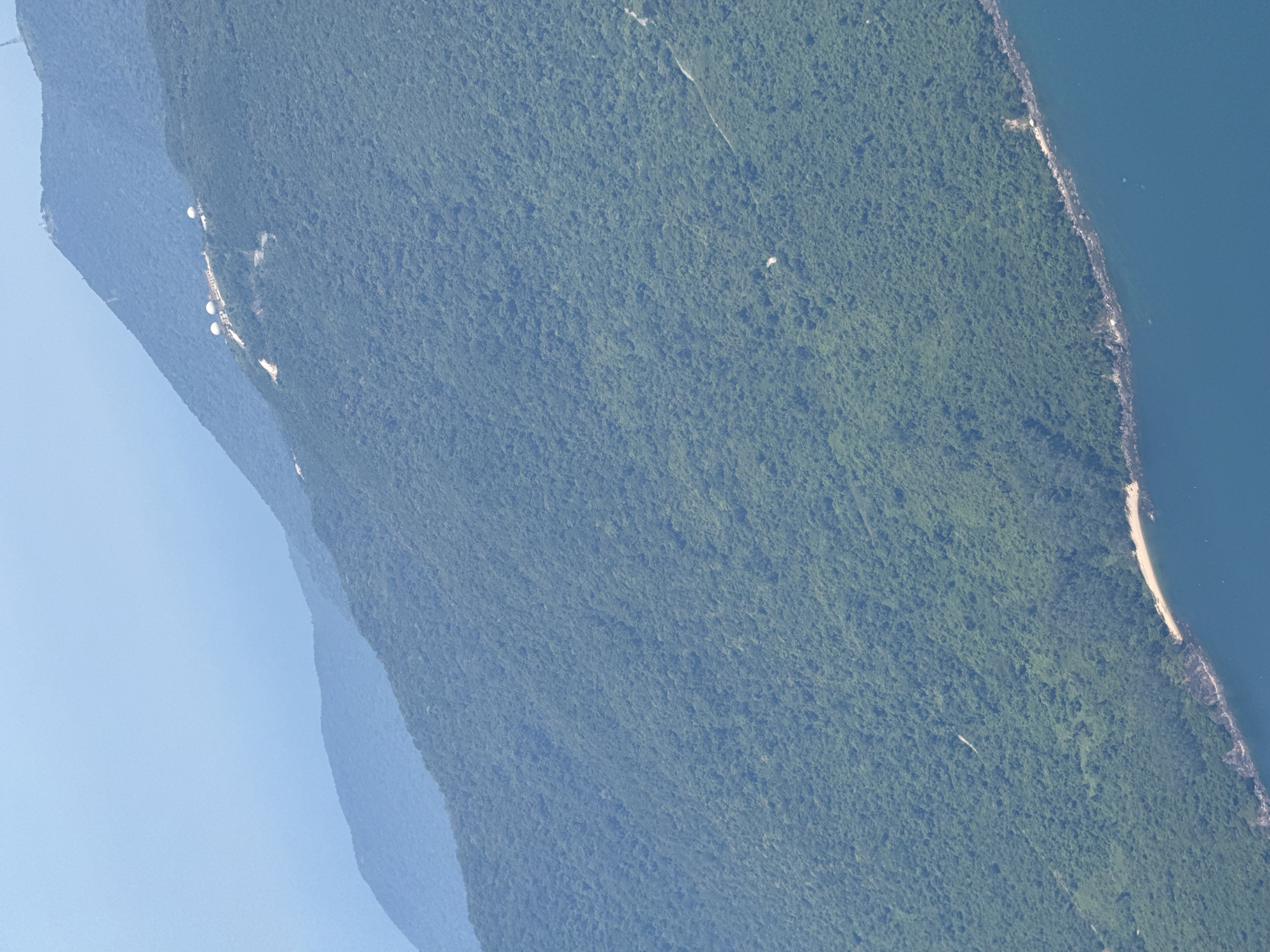 This aerial image depicts a mountainous, heavily forested area. The vegetation appears dense and green. To the right, the forest meets a body of water. There's evidence of a sandy shoreline along this coast. On the left, the land slopes downwards to a blue sky. A distinct structure, possibly a series of buildings and spherical tanks, is visible near the mountainside. The general location appears to be a coastal region with significant forestation. The features such as steep mountains, dense forests, beaches along a body of water and built structures suggest that this location might be the area around Đà Nẵng, Vietnam.