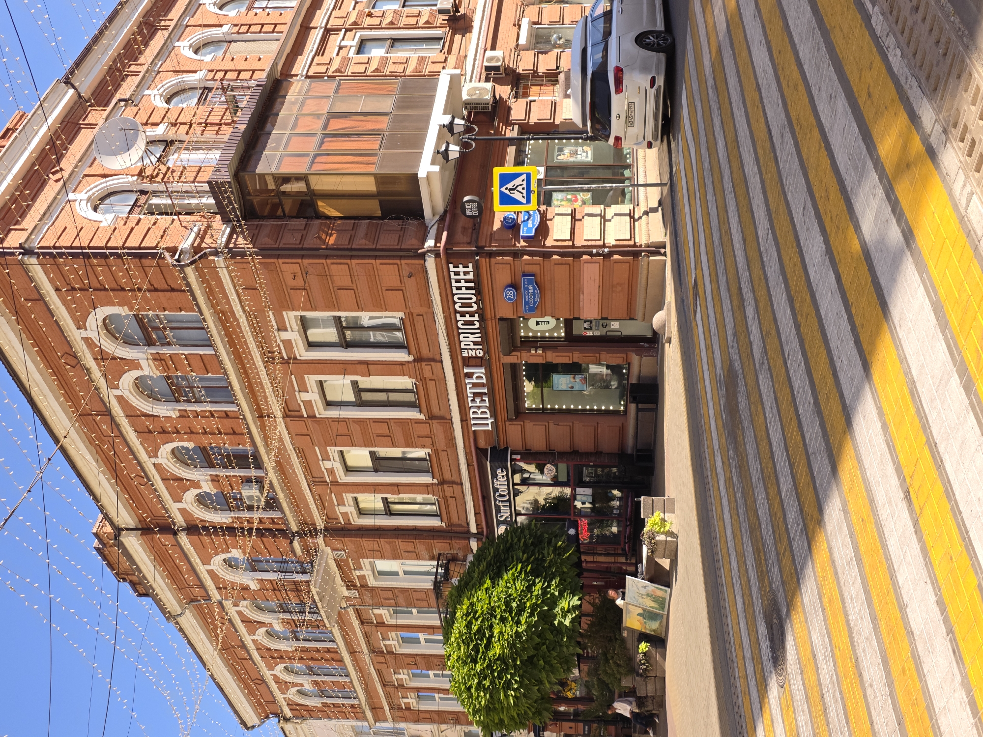 The image depicts a street view in the city of Rostov-on-Don, Russia, on a sunny day. In the foreground, a pedestrian crossing with yellow and gray stripes is visible. A white Kia minivan is parked on the right side of the street, facing away from the camera.
The background features a multi-story brick building with white trim and arched windows. The building's facade is decorated with strings of small white lights, suggesting a festive atmosphere or recent decoration. On the ground floor of the building, there are several storefronts, including "Surf Coffee" and a shop labeled with "ЦВЕТЫ" (Flowers). A large, round, green tree stands in front of the building, providing shade. Two individuals are seated on a bench near the tree, and several framed artworks are displayed on the ground.
Street signs and traffic signals are also present, indicating a typical urban intersection. The sky is clear and blue. The overall scene is a vibrant snapshot of a city street, likely a commercial area, under bright daylight.