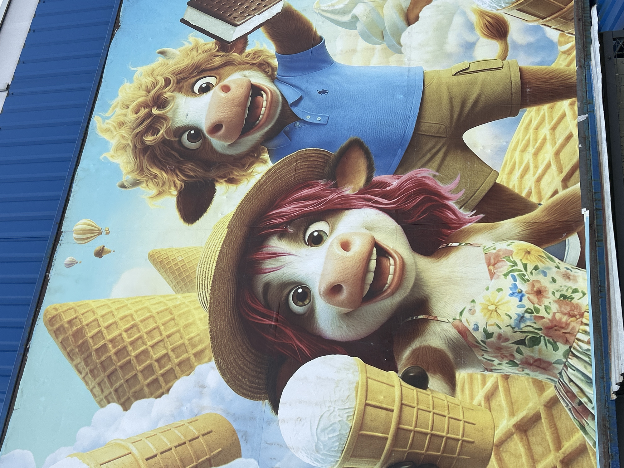This is a mural painted on the exterior wall of a building in Bila Tserkva, Ukraine. The mural depicts two anthropomorphic cows enjoying ice cream. The cow on the left wears a straw hat and a floral sundress, holding a large ice cream cone. The cow on the right wears a blue polo shirt and khaki shorts, holding an ice cream sandwich. They are surrounded by oversized ice cream cones, hot air balloons in the sky, and a waffle-patterned background. The mural is vibrant and whimsical, conveying a sense of fun and summer enjoyment.