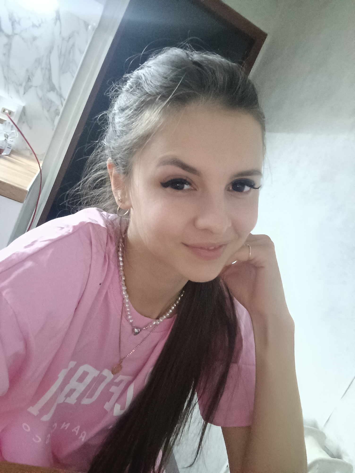 A young woman is posing for a selfie indoors. She has long dark hair pulled back, wears a pink t-shirt, and has two necklaces: one a pearl necklace and the other a gold chain with a pendant. Her left hand is resting on her face, which has makeup, including eyeliner and eyelashes. The background includes a doorway with white molding and a white and gray patterned wall. The location is Unknown, Unknown.
