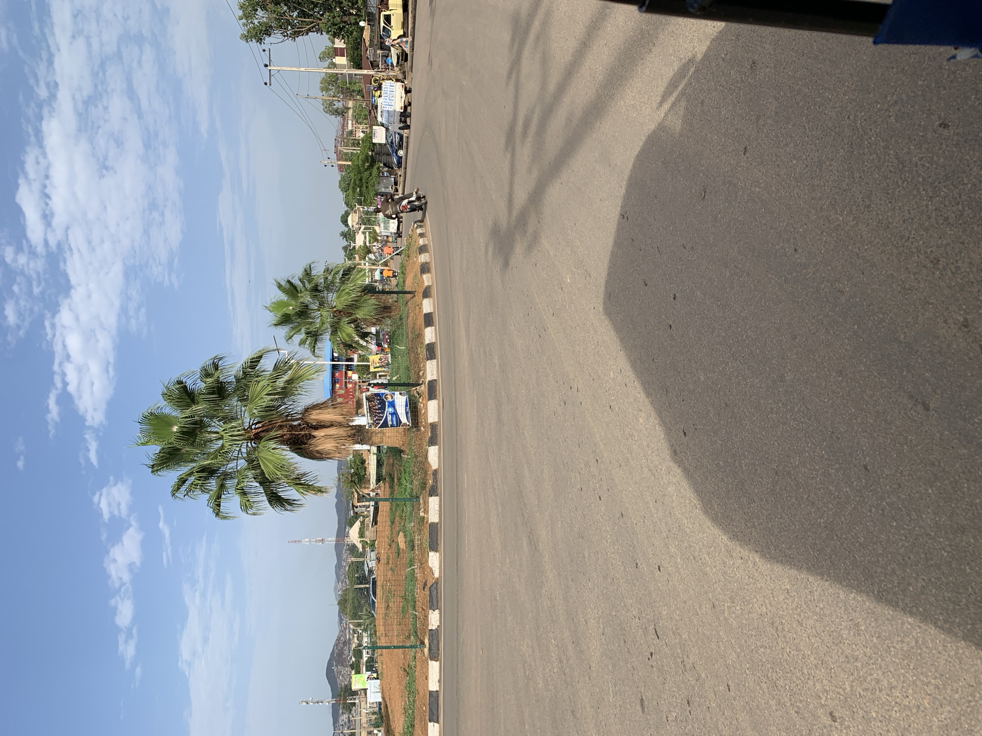 The image shows a road in Rafin Par, Nigeria. The road is asphalt with a black and white striped curb on one side. Along the curb, there are palm trees and various signs. In the background, buildings and utility poles are visible. The sky is blue with scattered clouds. A shadow is cast across a portion of the road.