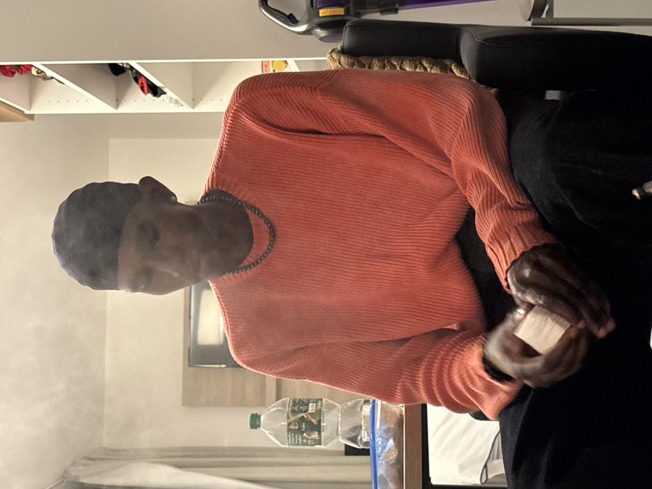 The image depicts a single individual seated indoors within what appears to be a residential room or hotel room located in Paris, France. The person, a male with dark skin, is shown from the waist up, oriented slightly to the right with his head tilted downwards. He wears a dark head covering, a light coral-orange ribbed long-sleeved sweater, dark pants, and a dark beaded necklace, with an earring visible in his left ear. A plume of white vapor or smoke is visible emanating from his mouth. His hands are clasped, holding a stack of small, light-colored rectangular objects.
To the left of the subject, on a light-colored surface, is a partially consumed large plastic bottle of water with a green cap and a label featuring a male figure. Adjacent to it is a clear plastic container holding brown food items. Behind these objects, a wall-mounted television screen, turned off, is visible above a light-colored wooden panel.
To the right of the subject, a white shelving unit with multiple open shelves holds various folded items, including garments or accessories in red, black, and white. A purple and yellow/gold upright vacuum cleaner is partially visible beside the dark, padded chair occupied by the individual. The room is illuminated by artificial light.