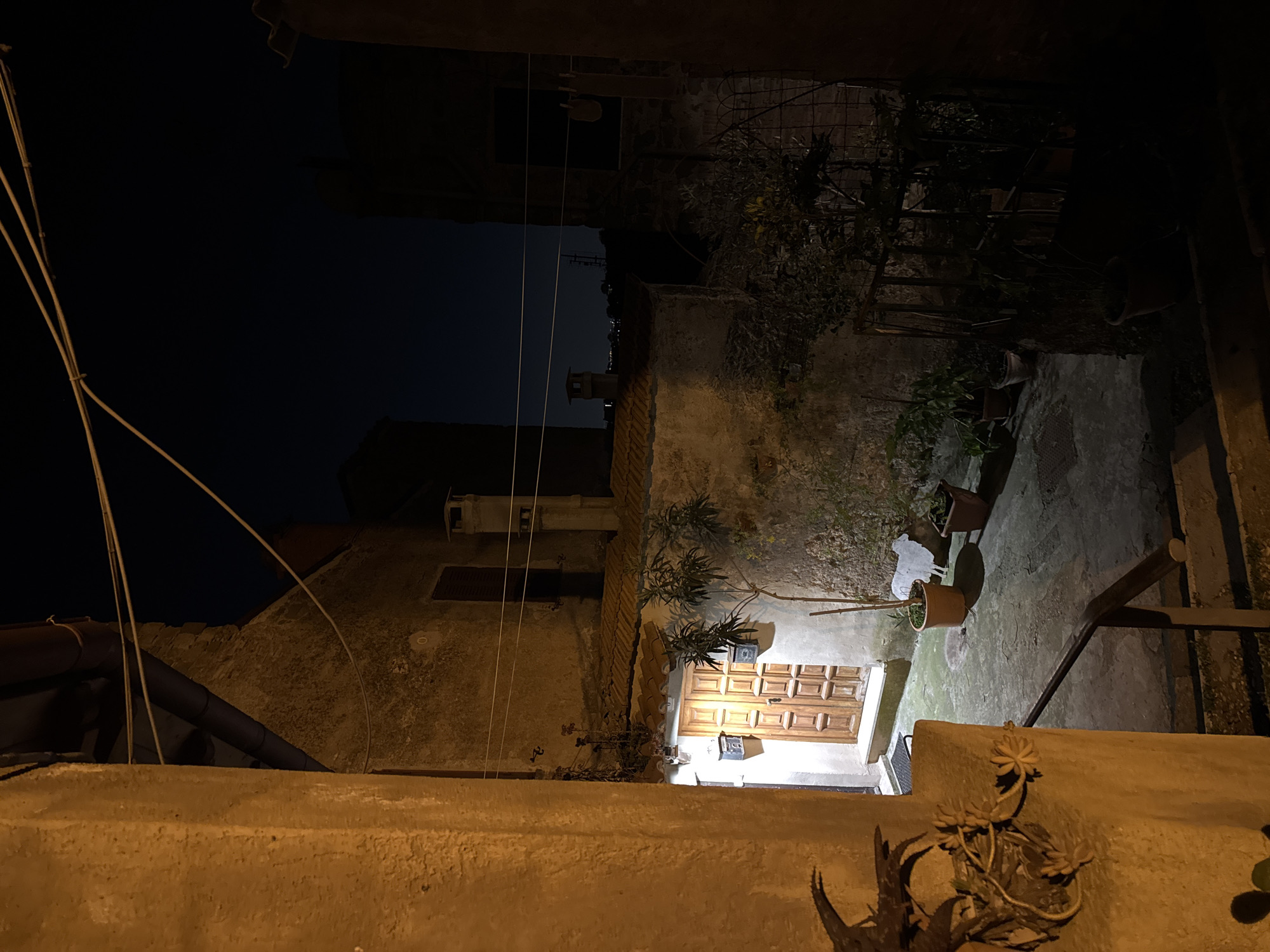 The image captures a night scene in Castelnuovo di Porto, Italy. A narrow, walled courtyard is illuminated by light emanating from a doorway. The door is wooden and framed by light fixtures. Potted plants are visible on the courtyard floor. The surrounding walls are weathered, and wires stretch across the open space above. Above the walls, a building facade with a window and additional architectural features is apparent. The sky above is dark, with faint hints of distant city lights. The viewpoint is from an elevated position, looking down into the courtyard. In the foreground, a concrete wall with some foliage growing on it.