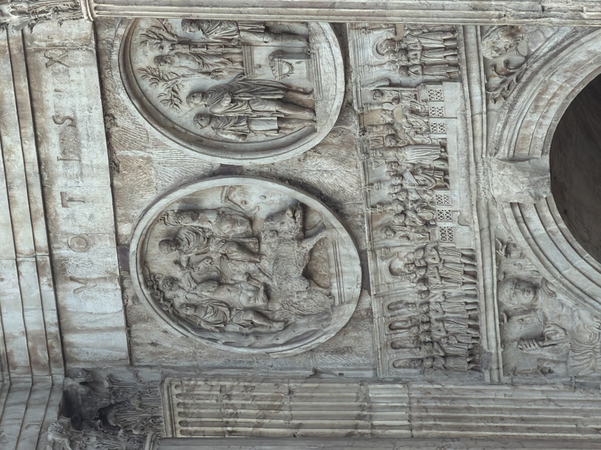 This is a close-up, low-angle, exterior shot of a weathered stone relief sculpture on an ancient architectural structure. The sculpture depicts a frieze of figures and animals. At the top, there are carved Roman numerals "VOTIS X", possibly indicating a decennial vote or offering. Below this inscription, two circular reliefs are visible. The left circular relief shows a scene with several figures, some on horseback, and a boar. The right circular relief features a group of standing figures, one holding a vessel, and another figure near a pillar. Beneath these circular reliefs, a long, horizontal frieze depicts a procession of numerous standing figures, including adults and children, in draped garments, with architectural elements in the background. Further down, a large, arched section of the structure is visible, with more relief sculptures of figures, some appearing to recline or be seated. The stone is heavily textured, showing signs of age and erosion, with variations in color from light grey to brown. The overall impression is one of historical grandeur and significant age.