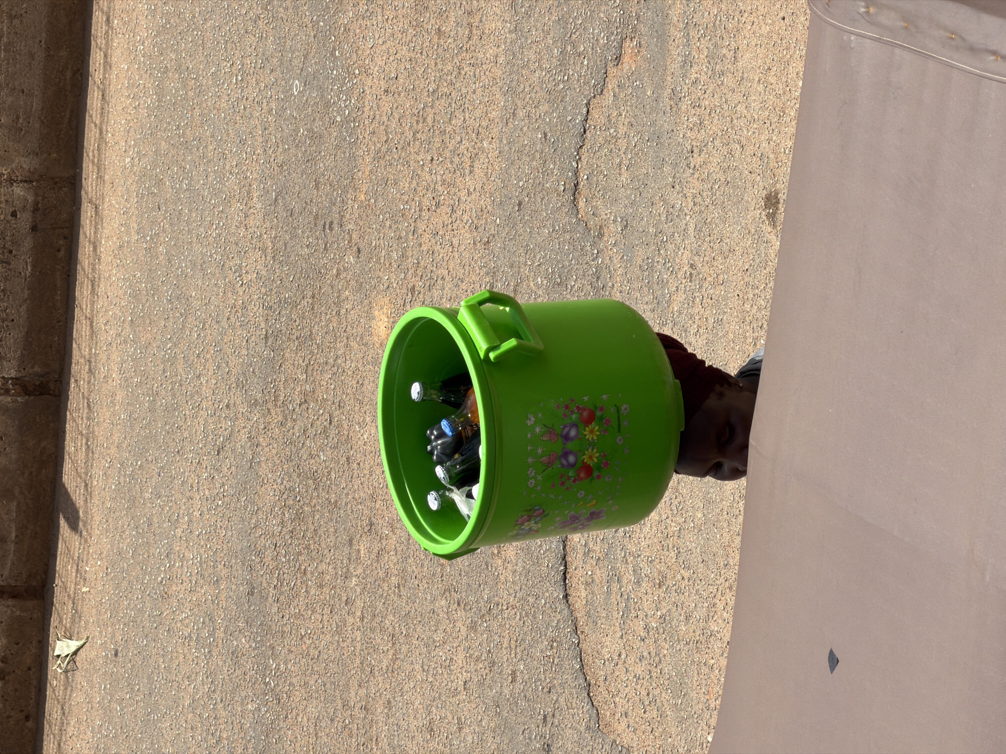 A person carries a bright green plastic bucket filled with bottled beverages on their head, walking on a dusty asphalt road in Bukuru, Nigeria. The bucket is decorated with colorful floral and butterfly motifs. The person's face is partially visible, showing dark skin and eyes. They are wearing a dark head wrap. The background consists of the textured asphalt road and a brick wall. The overall scene depicts a street vendor in a Nigerian city.