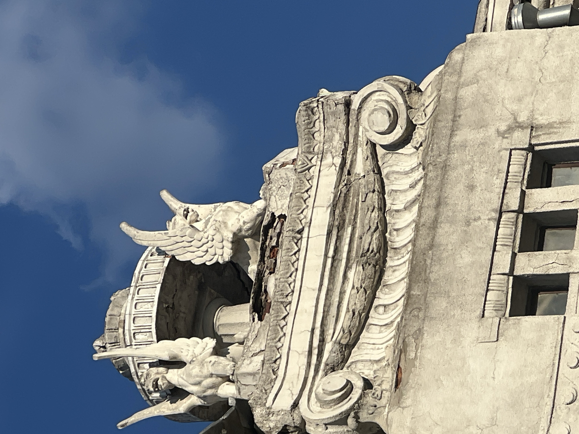 This is a close-up, low-angle, outdoor, daylight shot of the dilapidated upper facade of the Kharkiv National Academic Opera and Ballet Theatre, located in Kharkiv, Ukraine. The focus is on architectural details and decorative elements, with a clear blue sky with scattered clouds in the background.
Prominently featured are two winged, mythical creature sculptures, likely griffins, made of stone or plaster. The sculpture on the left is positioned slightly further back and angled towards the right, while the one on the right is closer to the viewer and faces left. Both have discernible heads with open mouths, large wings, and avian or feline features. They appear weathered and in need of repair.
Below the sculptures, a section of the building's cornice exhibits significant deterioration. Cracks are visible, and chunks of the facade have fallen away, revealing red brickwork underneath. The cornice itself is ornately decorated with scrolling volutes on either side, a draped festoon pattern, and a geometric frieze. The material appears to be weathered stone or stucco.
Further down, a portion of the building's wall is visible, featuring recessed window frames or decorative panels with vertical striations. A metal pipe or vent extends from the upper right side of the image, likely part of the building's utility system. The overall impression is one of historical grandeur marred by decay and the passage of time. No people are visible in the image.